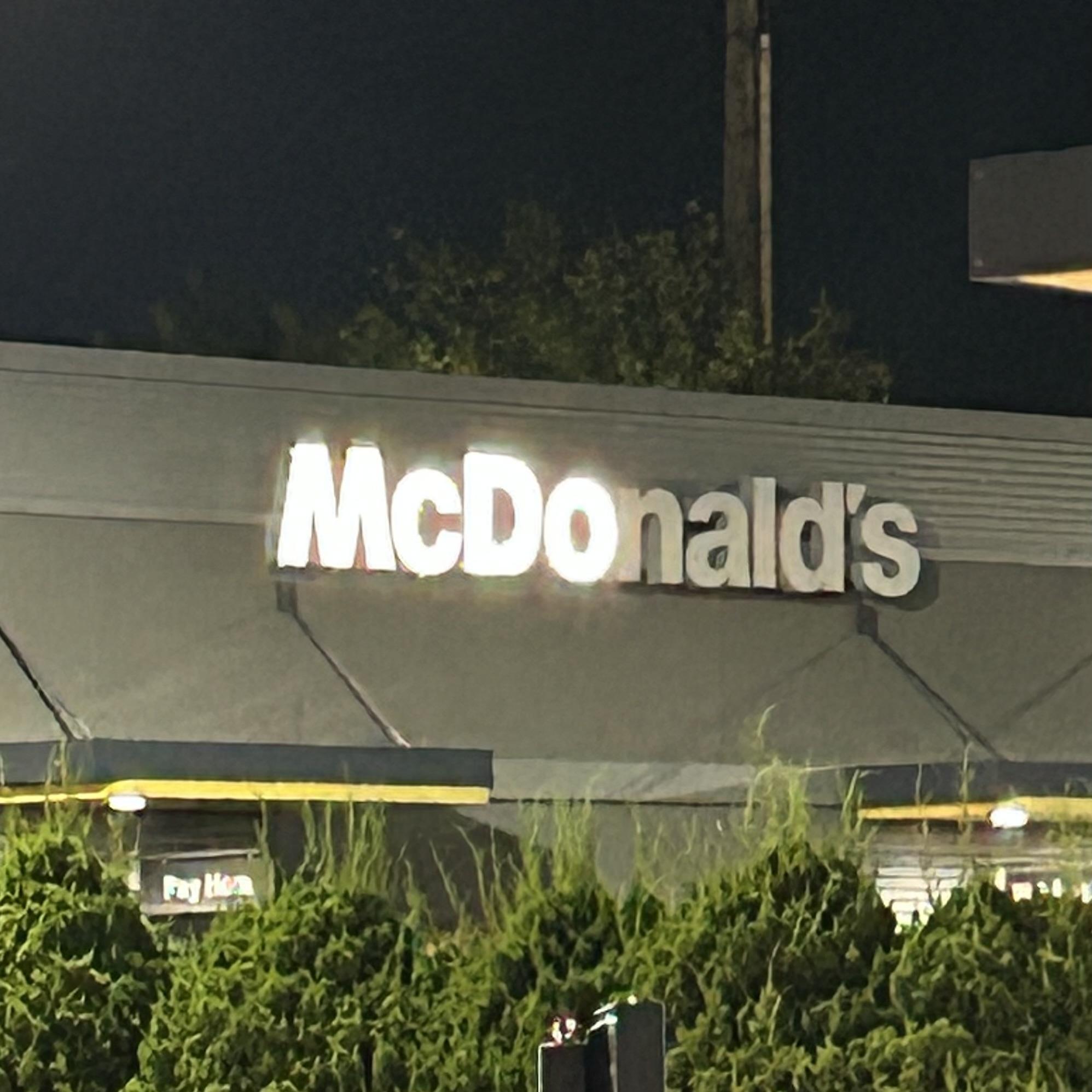 Nighttime photo of a McDonald’s exterior, centered on the large illuminated “McDonald’s” sign mounted high on a gray building facade. Only the first four letters are illuminated, spelling “McDo.” Dark trees and a utility pole rise behind the roofline, while dense green shrubs fill the foreground. Parts of the restaurant’s yellow-trimmed awnings and windows are visible near the bottom.