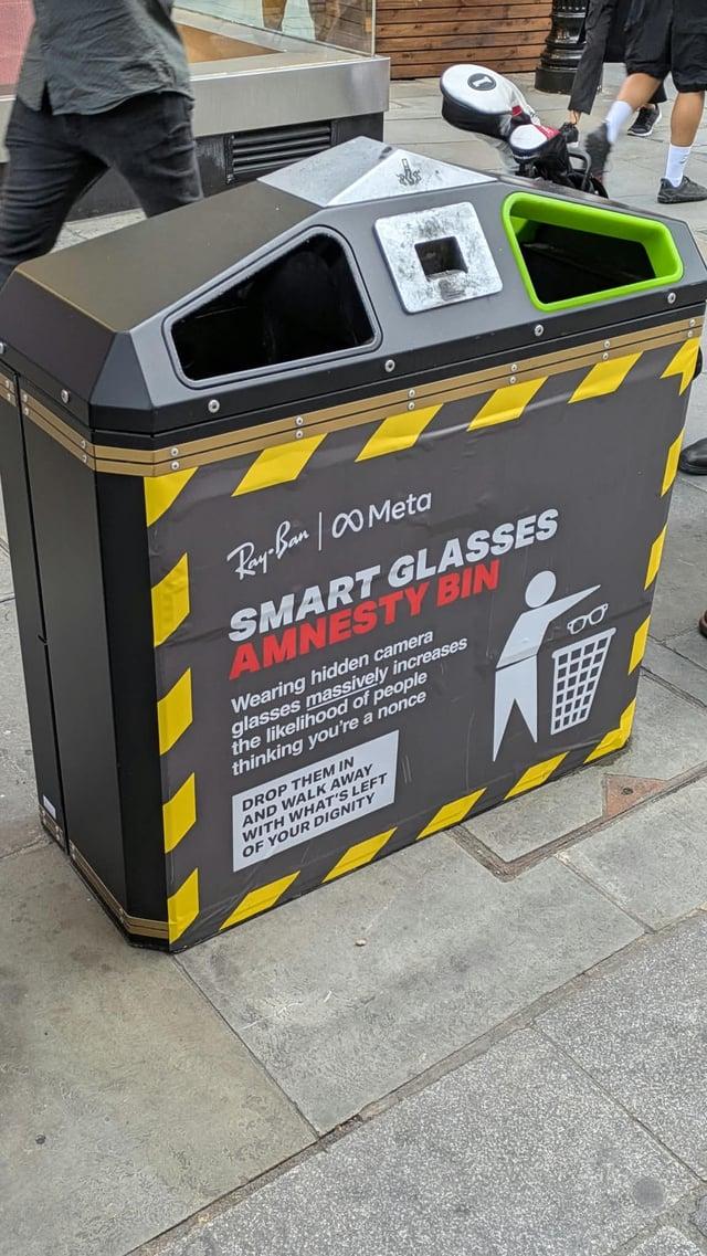 A bin with some ad busting layover stuck to it. Under the ray ban and Meta logo it reads "smart glasses amnesty bin" next to a pictogram of a person throwing the glasses into a bin.  Further text reads: Wearing hidden camera glasses massively increases the likelihood of people thinking you're a nonce DROP THEM IN AND WALK AWAY WITH WHAT'S LEFT OF YOUR DIGNITY