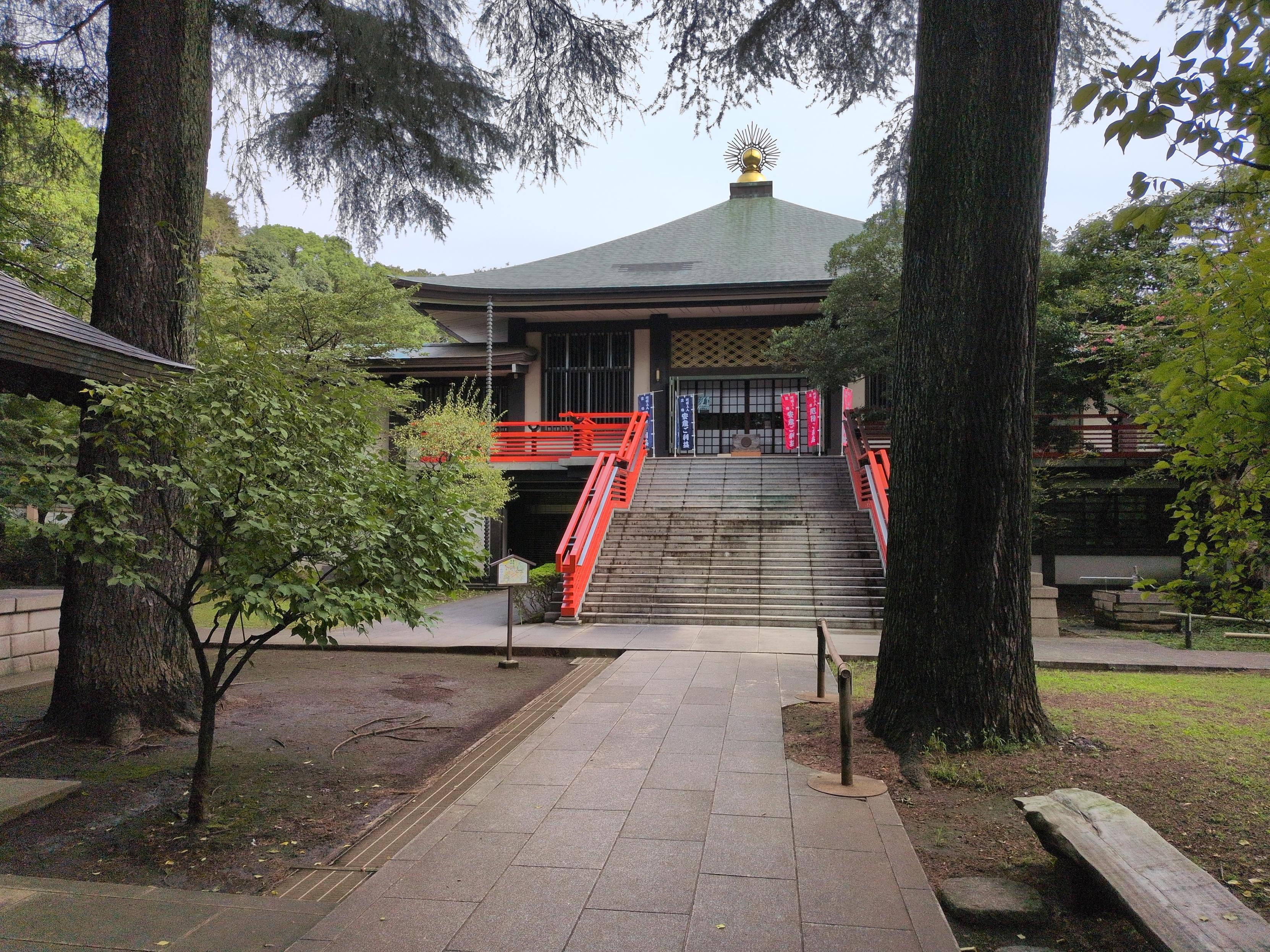 The main hall of this temple, seen from between two trees. There is a wide stairway with bright red railings, leading to a squat building with a green triangular roof and a golden ornament on top that looks like an spherical sun with needle-thin rays surrounding it. The sliding door entrance is currently closed, with tall blue and red cloth banners on either side with Japanese writing.