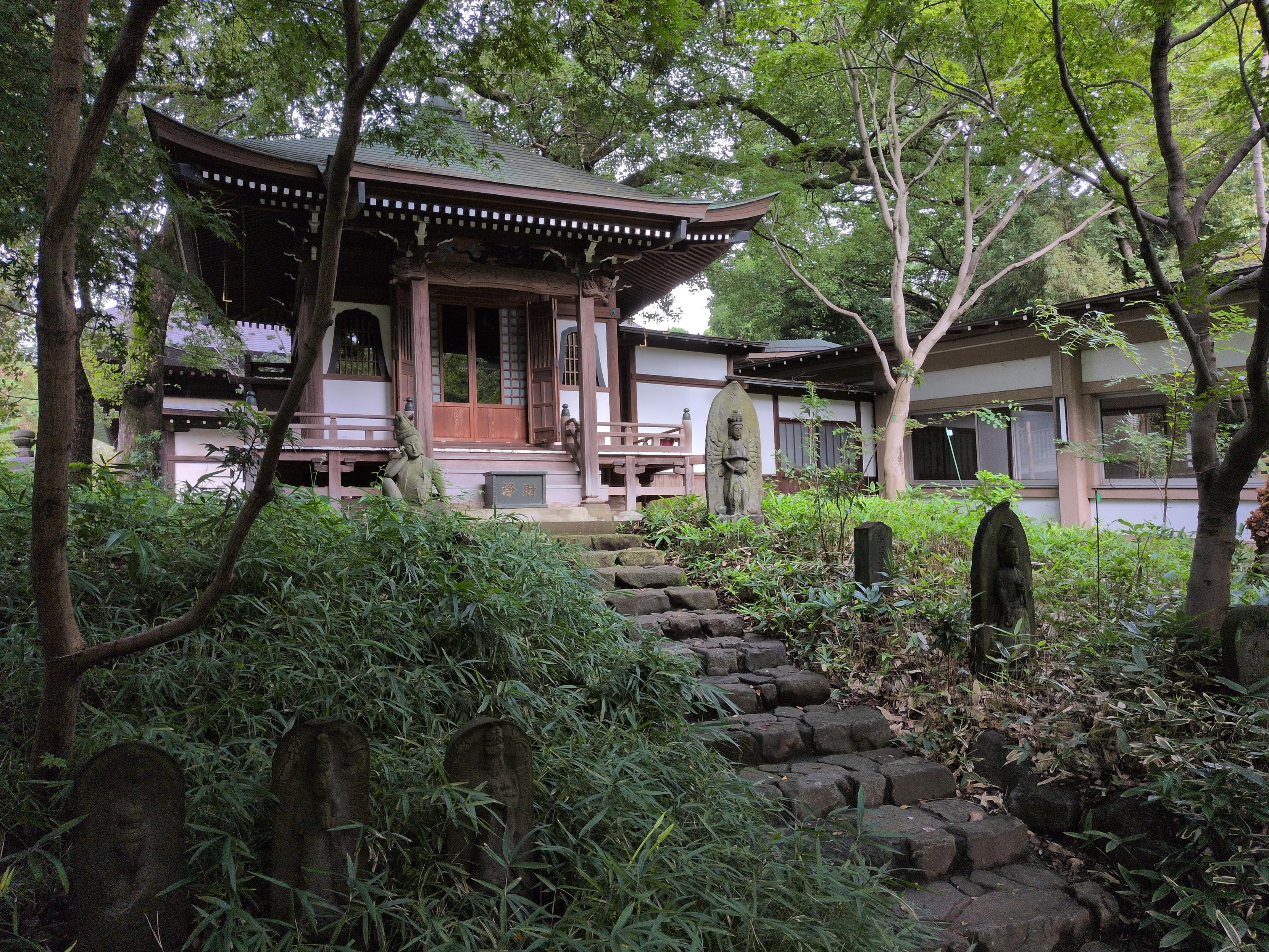 Stone steps cut through a lush, shaded area with trees and tall grass, which partially hides stone statues of bodhisattvas on either side. It leads to a small wooden building with a sweeping green-tile roof. The entrance has statues on either side, one seems to be a Buddha while the other is a standing figure with many arms.