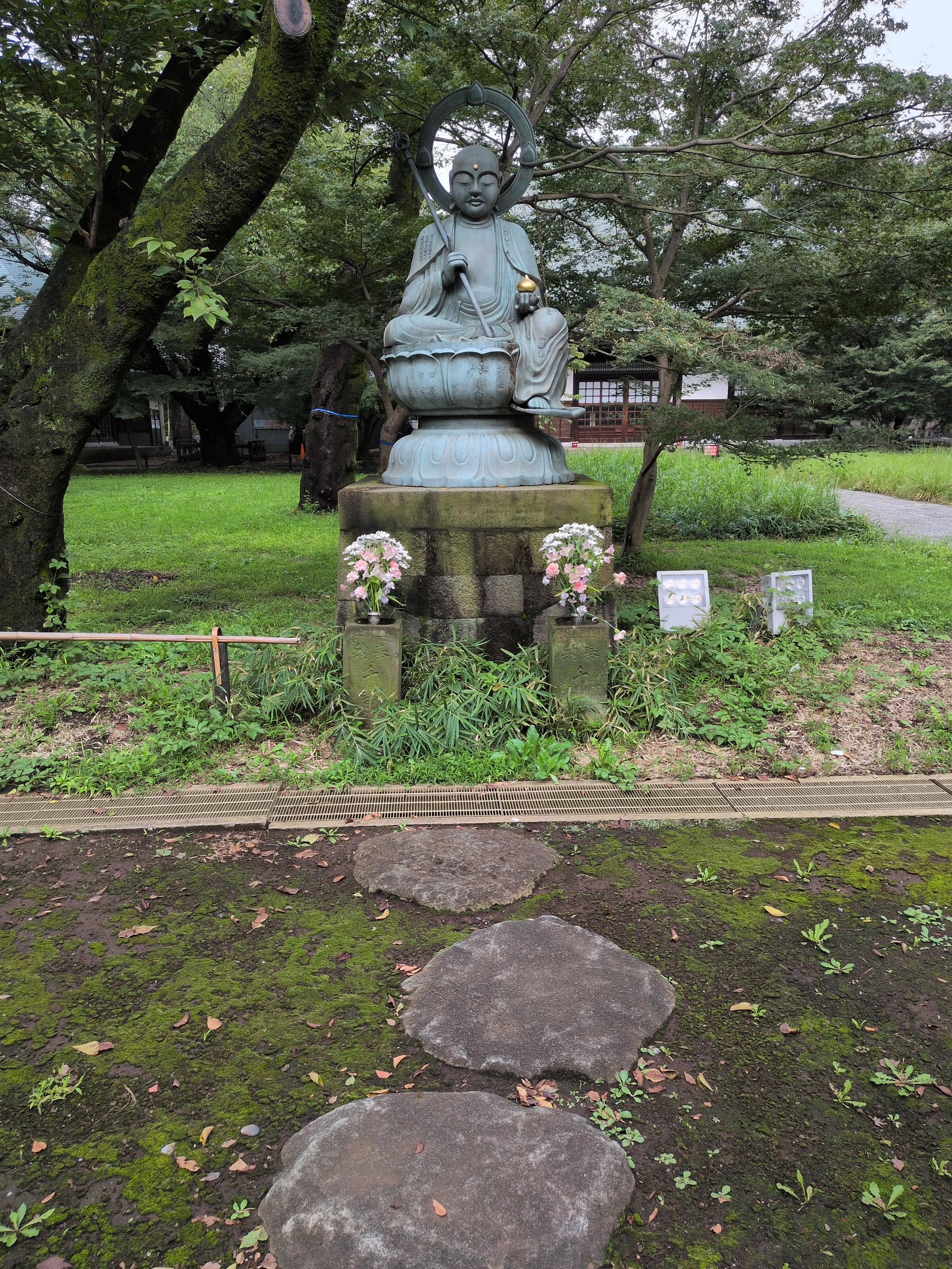 A green Buddha statue with gold accents sits on a stone base, decorated with two bouquets of pink and white flowers. A few round stepping stones lead up to it, with greenery and mossy barked trees behind.
