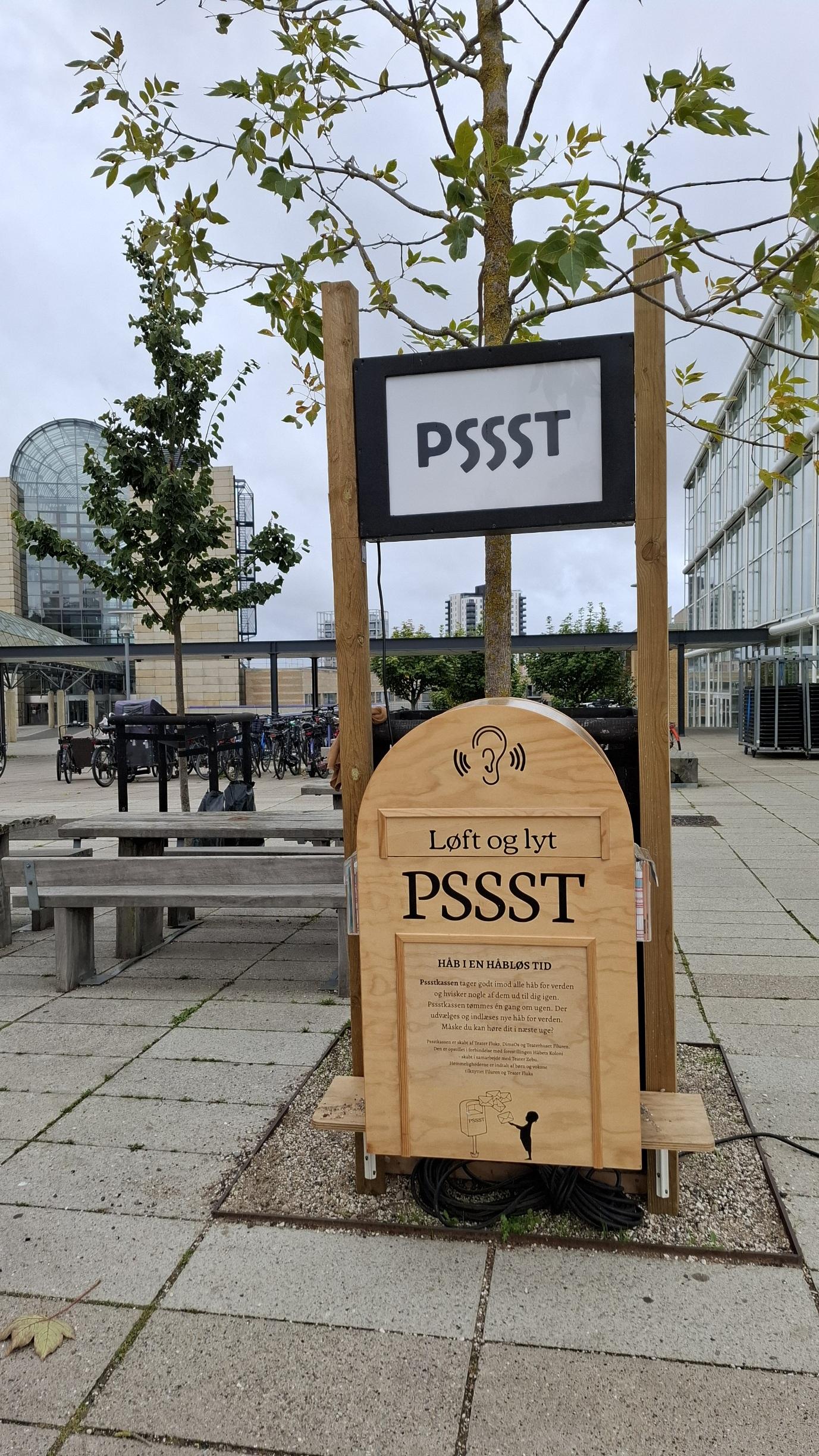 Short version: a wooden postbox outdoors. Fuller description: A light-brown wooden postbox on a pavement area. Behind it, a city building with glass arch, a tree, a bench, bikes parked in bike stands. Sky overcast. Above the postbox, a sign saying Pssst. On the postbox above the flap, an illustration of an ear with soundwaves showing it listening to something. On the flap, the words Lift and Listen. Written below, a description of the project as roughly translated and summed up by me in the toot, and credits for the various groups involved. This is in Aarhus, Denmark. The postbox was in concert hall park recently, I think it moves around.