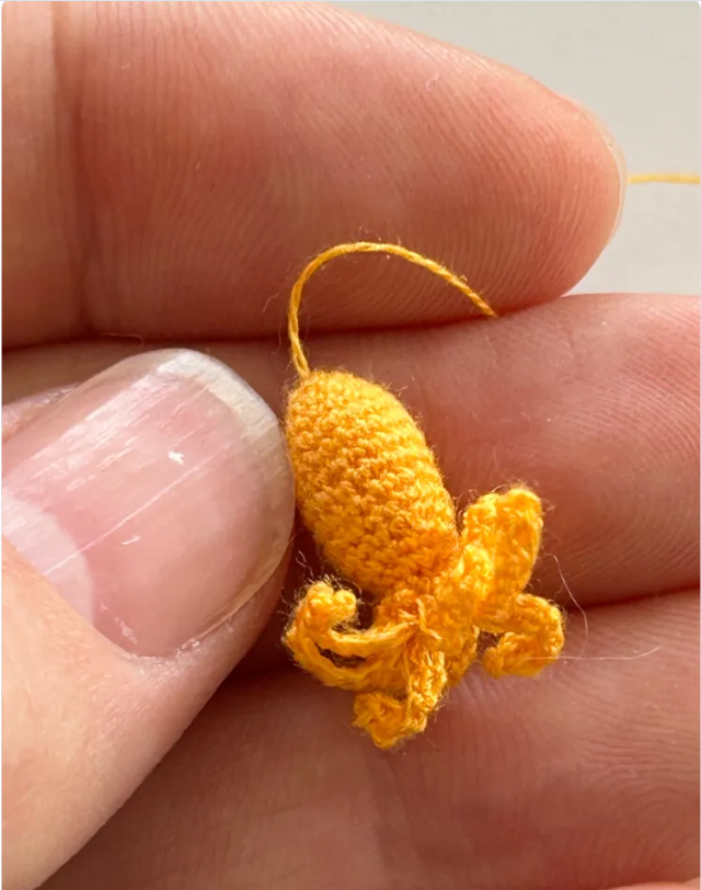 A photo of three fingers and a thumb holding a tiny yellow crocheted octopus. The octopus is smaller than the thumbnail visible in the picture.