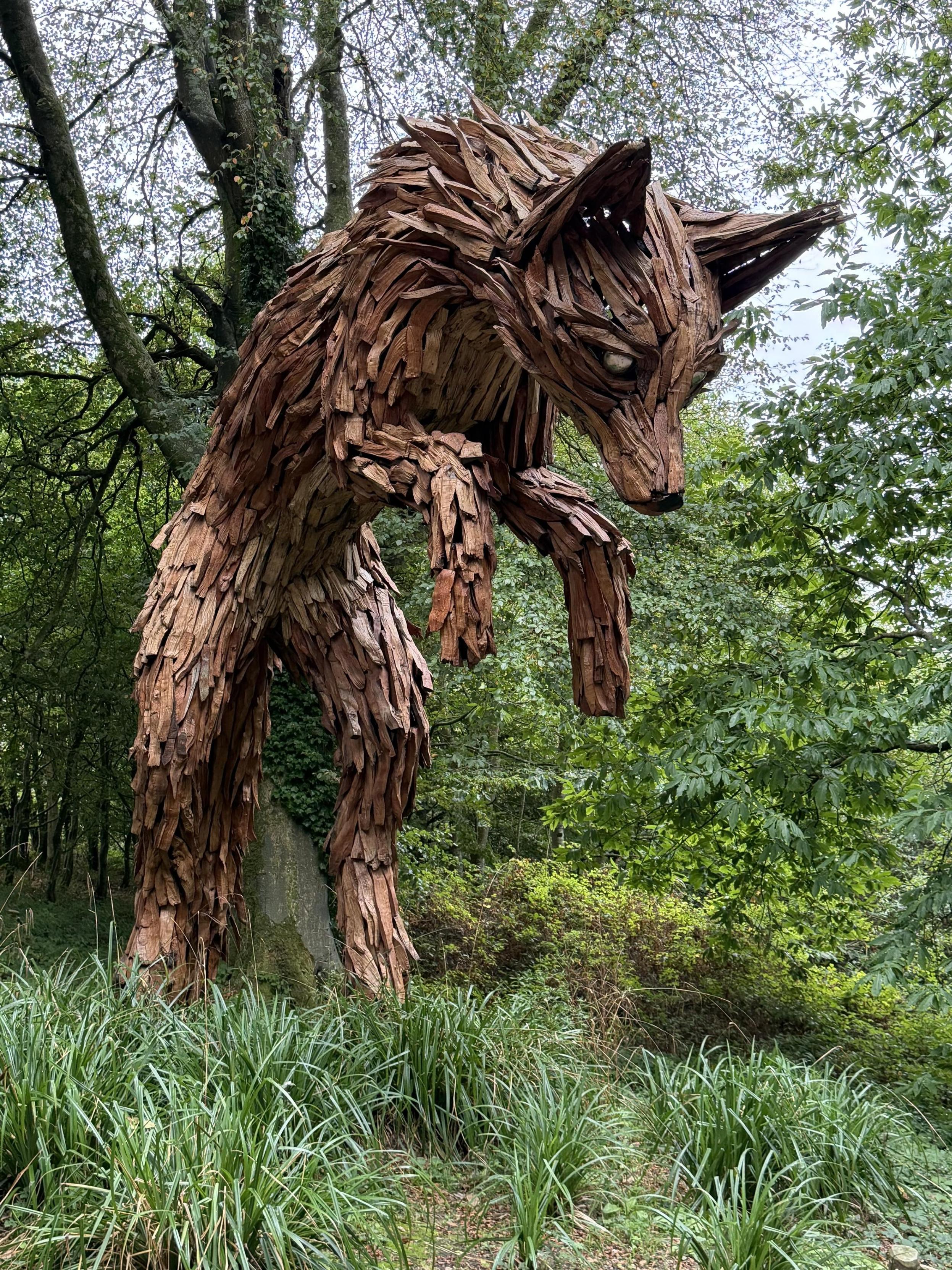 A large sculpture of a fox made from wooden pieces, positioned among trees and greenery in a forested area. The fox has a dynamic pose, appearing to leap or be in motion, with detailed features emphasizing its head and fur. It is looking down at the ground.