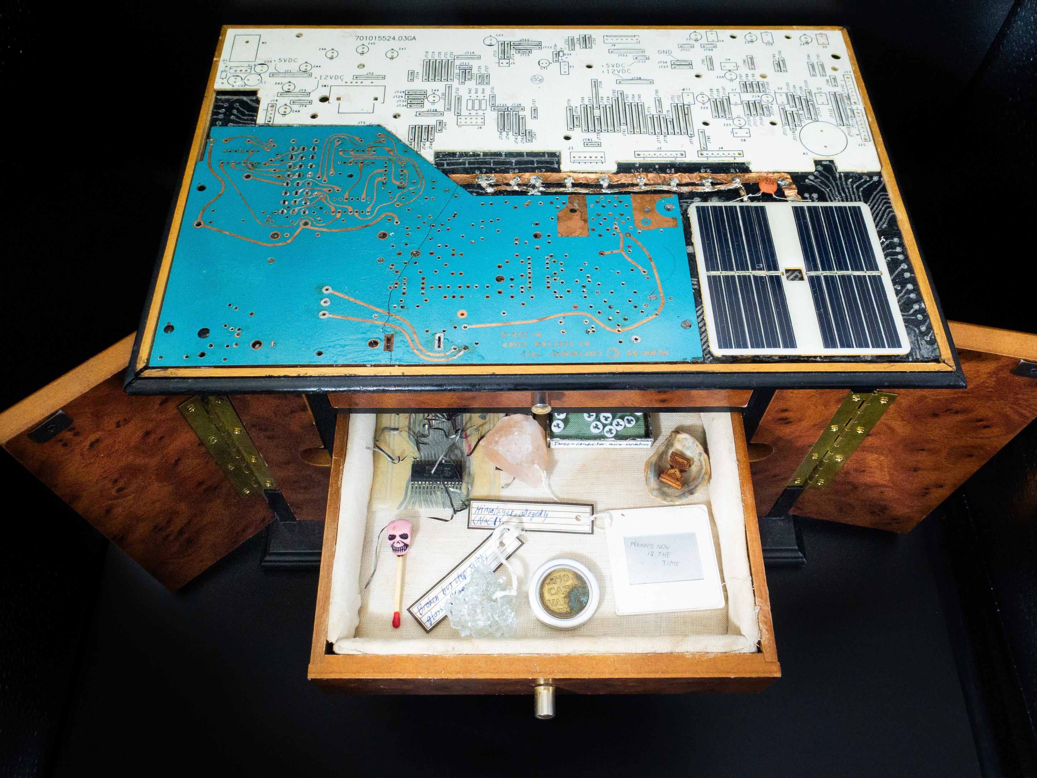 A tabletop cabinet is shown with one drawer hanging open. The drawer contains a number of curious objects, such as a tiny pink skull on a matchstick, a coin labeled "No Cash Value" in an aluminum shroud, a lump of Himalayan pink sea salt, and a tiny wooden hourglass in a seashell. Most artifacts are labeled with an old-looking label. The cabinet itself is strange, too; its top surface is made with a patchwork of vintage circuit boards, with some hand-drawn traces and unusual colors visible.