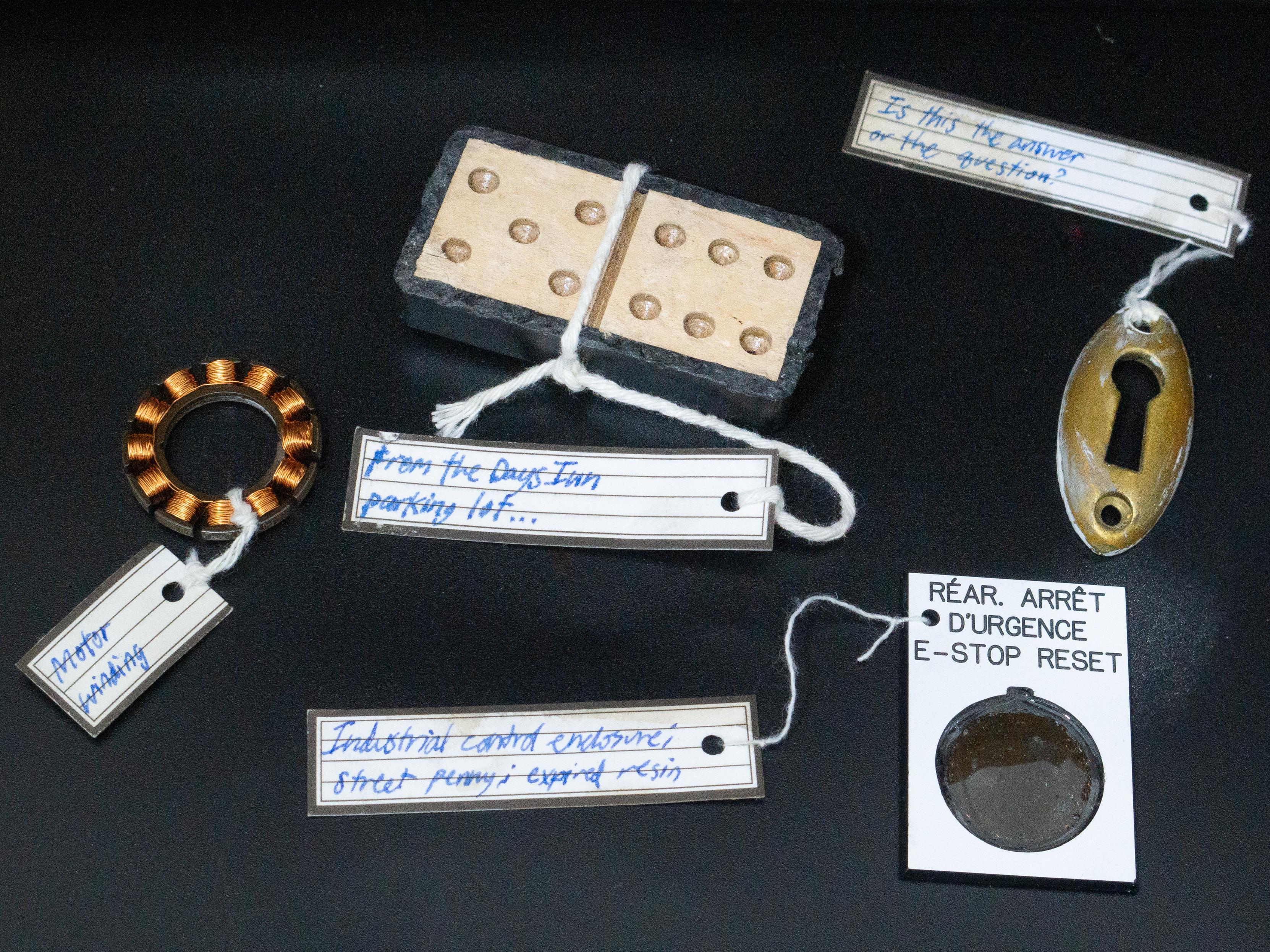 Four individual artifacts: A well-worn wooden domino, which looks almost handmade. Its label: "From the Days Inn parking lot..." A well-worn brass keyhole, labeled, "Is this the answer or the question?" Next to it is a dark, damaged penny In a plastic case which resembles a coin holder, but is a pushbutton legend from an industrial control enclosure reading "Réar arrêt d'urgence - E-stop reset." And finally, a curious little ring of bright copper windings, labeled "Motor winding."