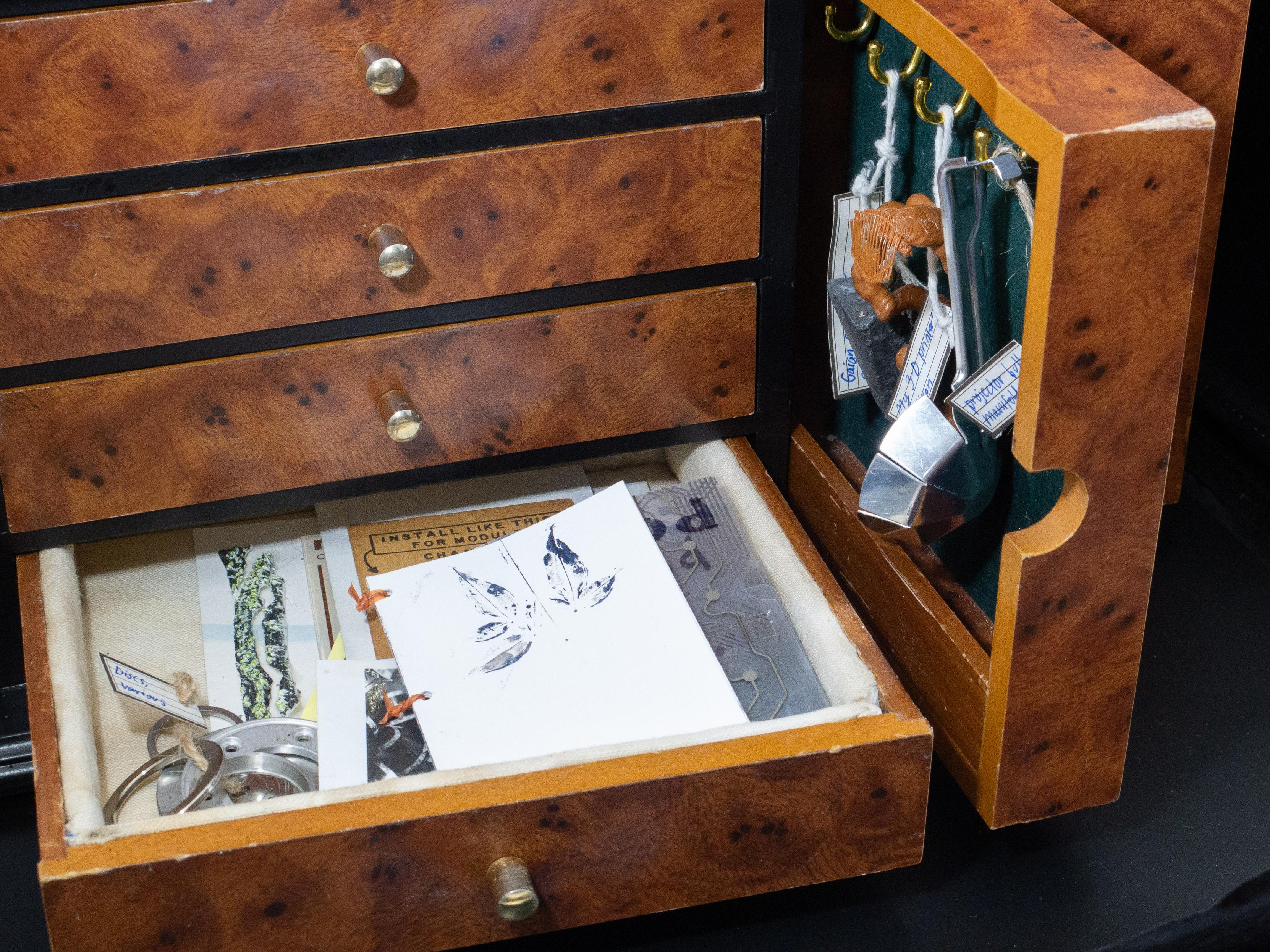 The front of the wunderkammer with a horizontal and a vertical drawer open. Hanging objects are a little tough to see, but they include some strange blobby orange plastic and a sort of metal scoop (actually a projector bulb shroud). The other drawer contains mostly paper goods, tiny zines and artworks, such as a gel-printed leaf.