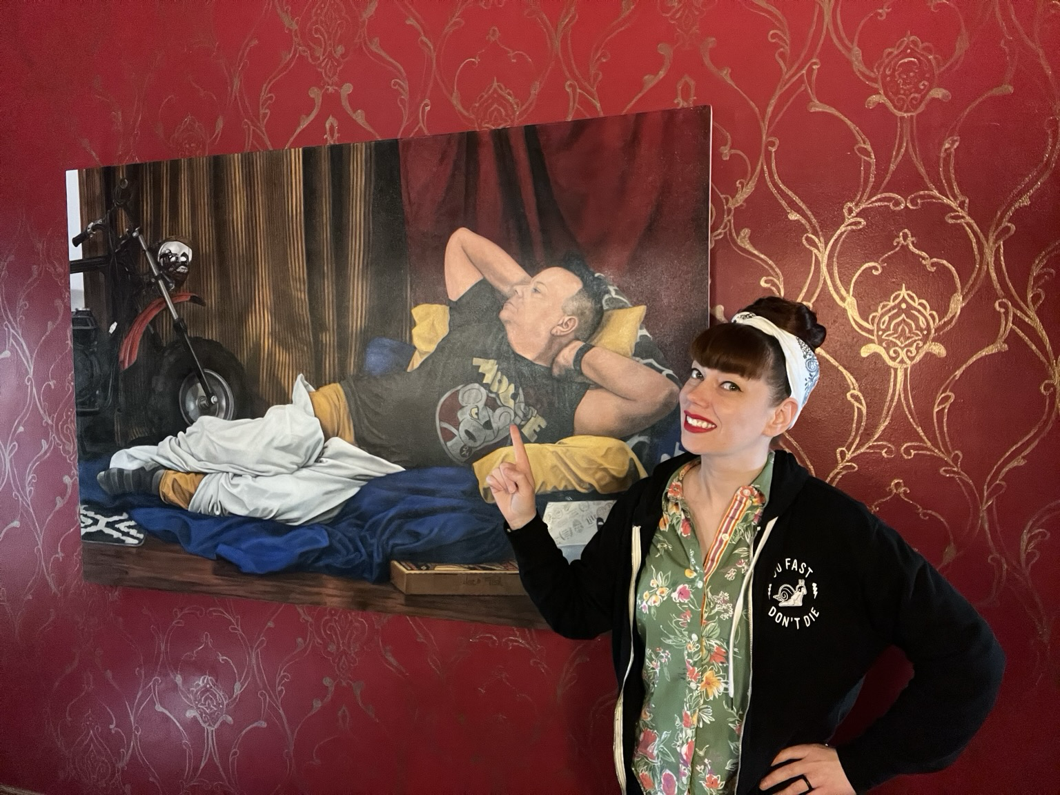 In this photo, a cheeseball of an artist stands in front of a 6’ wide oil painting titled Brodalisque with Minibike. She points toward the handsome model, who lounges on cushions while clad in a Danger Mouse t-shirt and draped modestly with white fabric.