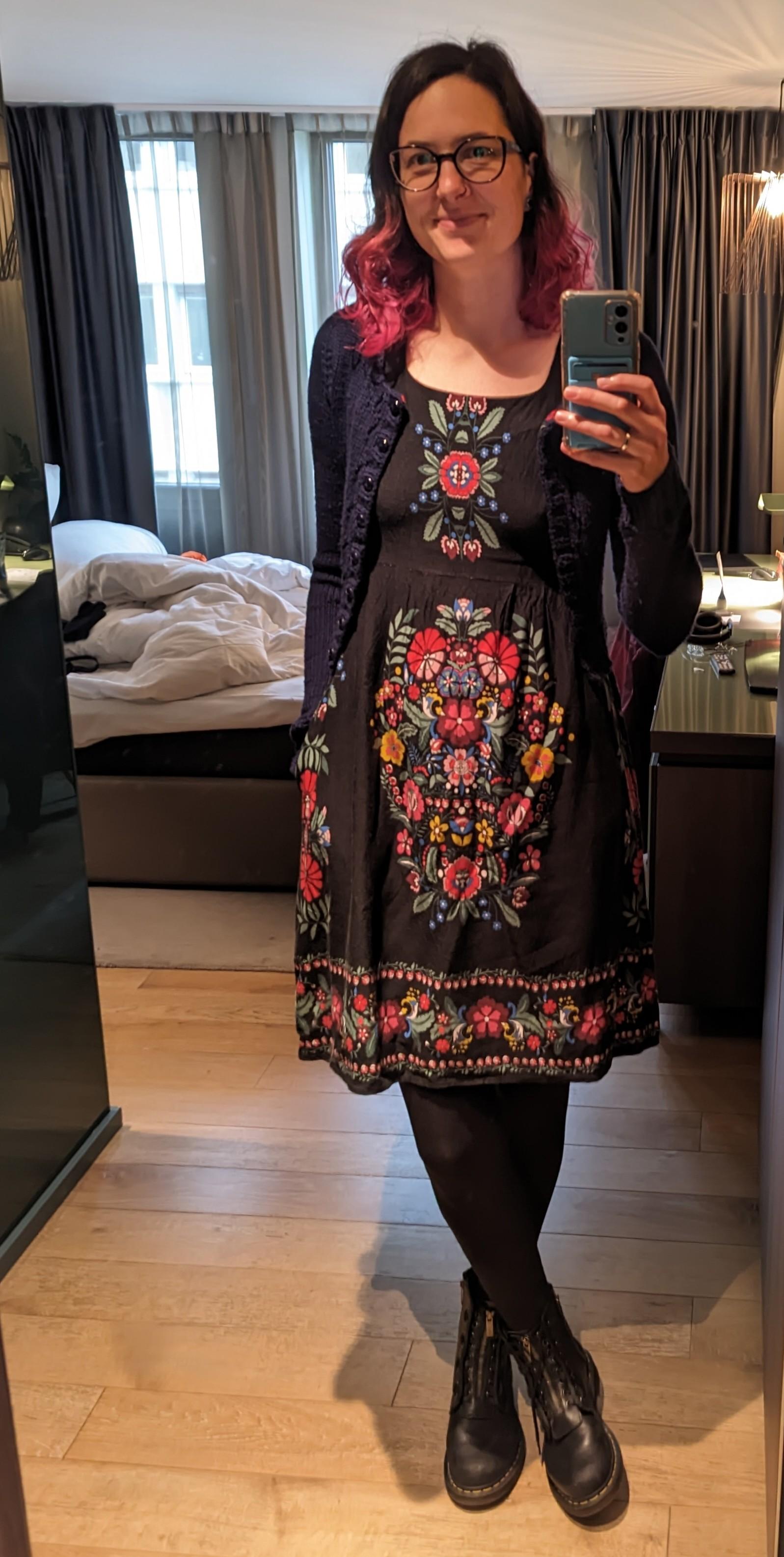 Unflattering hotel selfie of me wearing a black dress with a colorful flower pattern and a dark blue knitted cardigan.