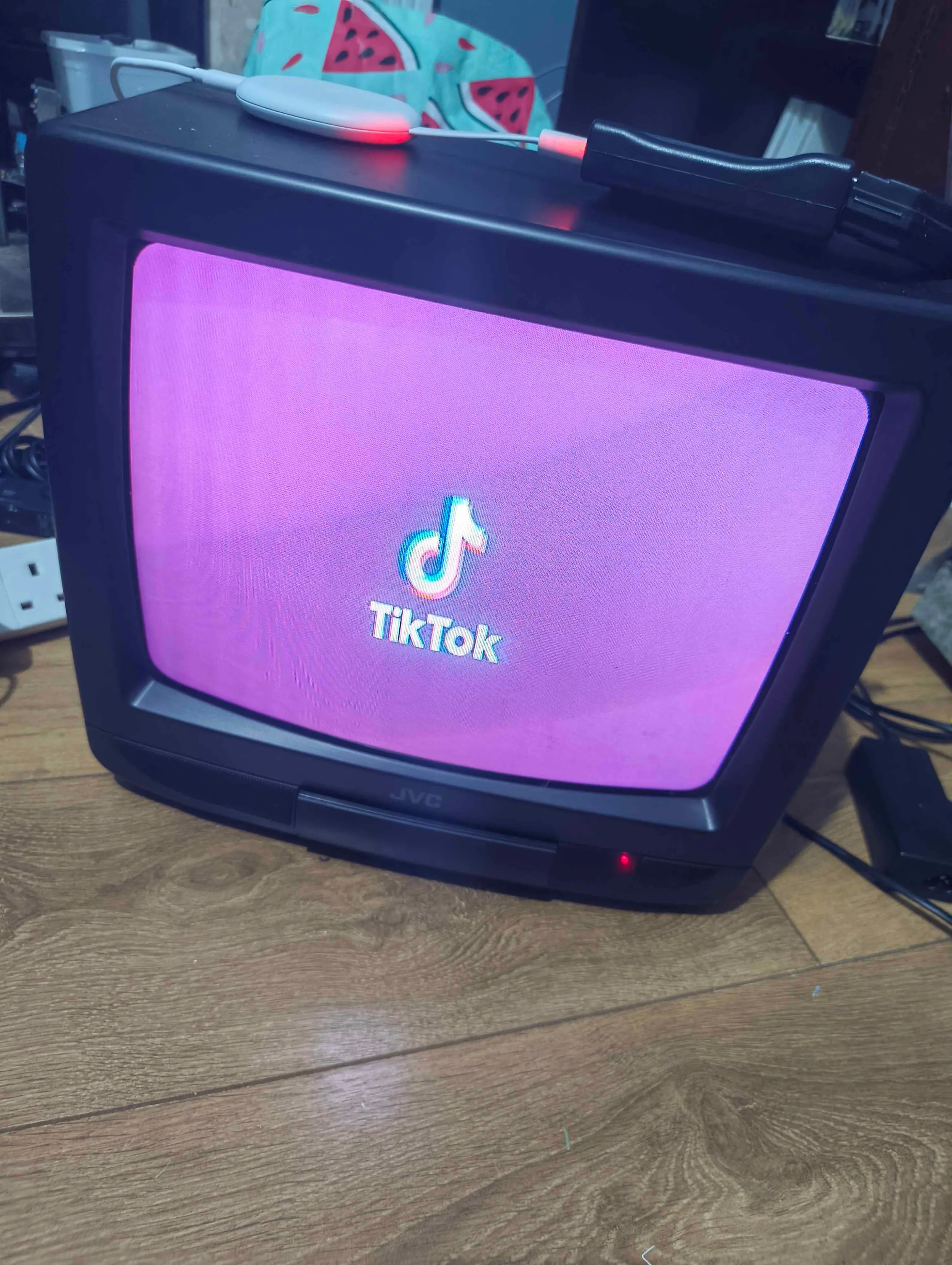 A Black Small JVC CRT TV Showing the Tiktok logo on the screen with a purple background, , the TV is on a wooden floor, with cables around it
