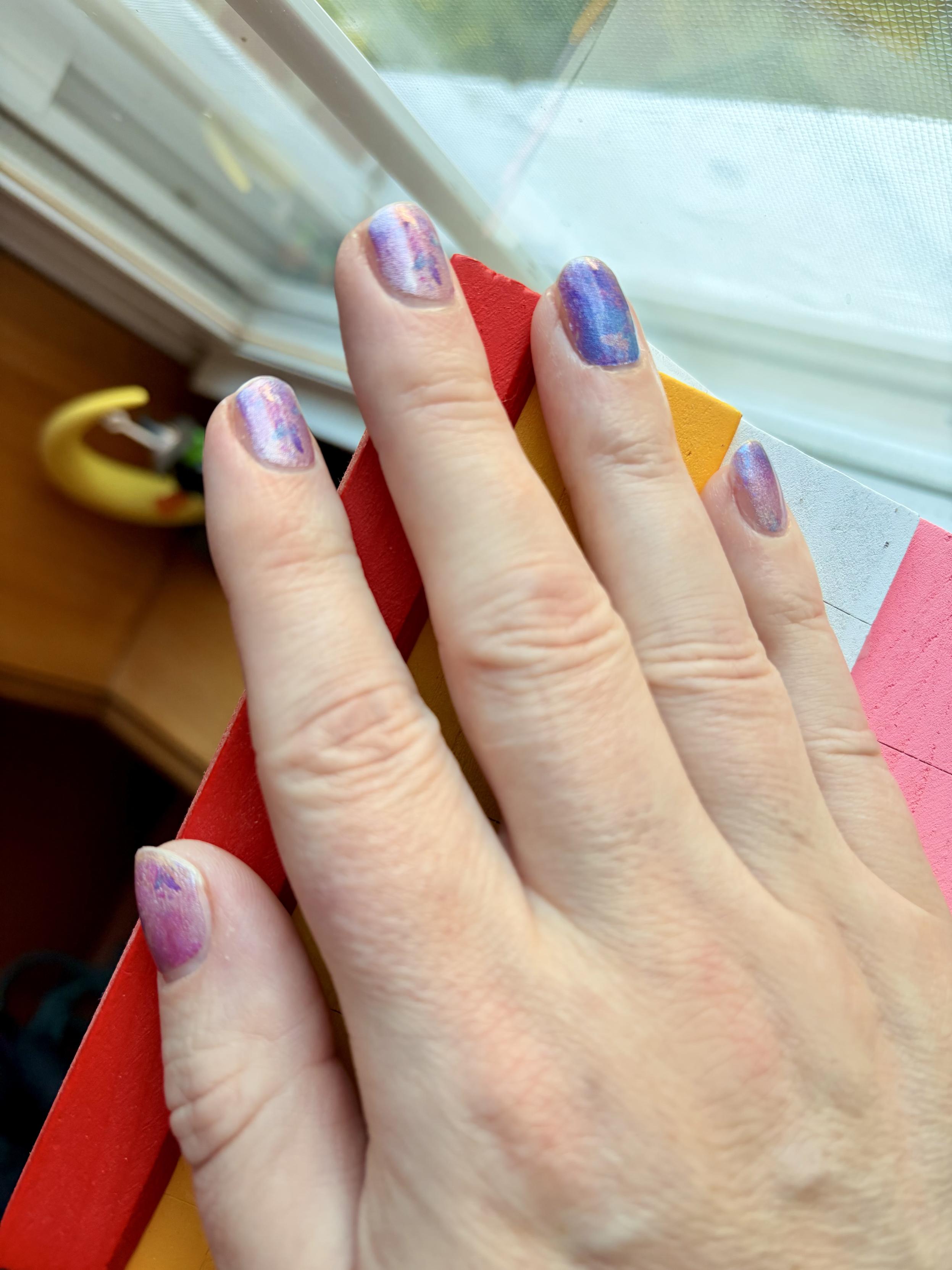 A slender hand with freshly decorated fingernails resting on a lesbian flag colored birdhouse perched on a window sill.  The nails are a pinky purple metallic color with butterflies, and the ring finger is a darker blue purple color.