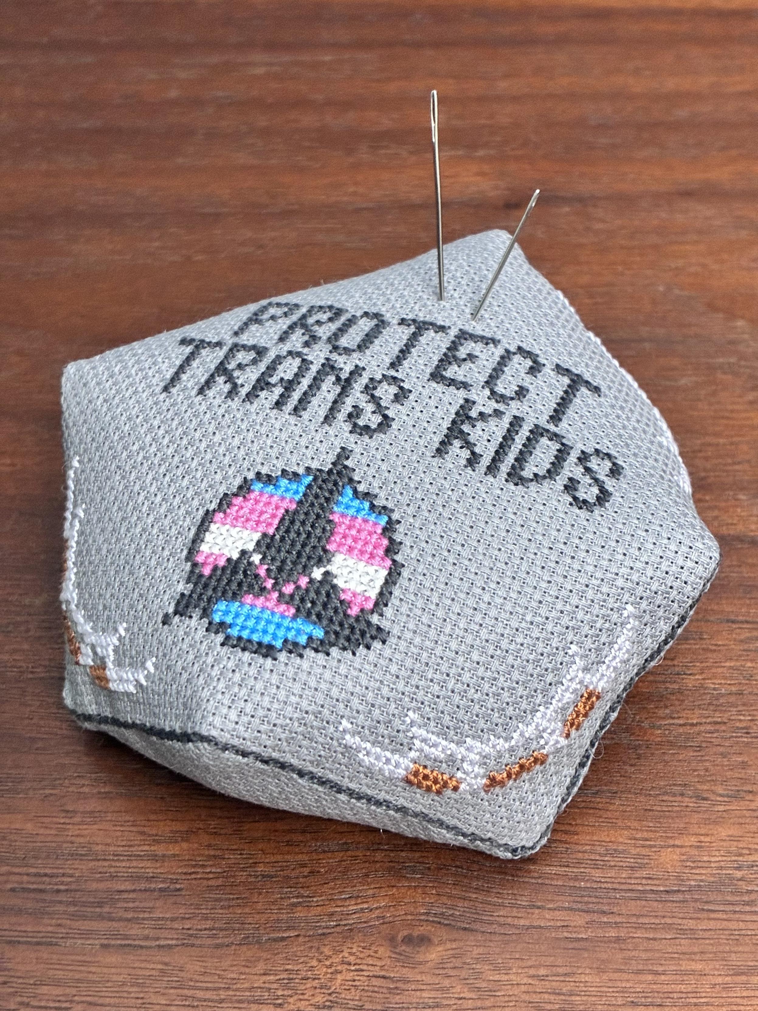 A biscornu, or weirdly- shaped octagonal, pincushion made from gray Aida and cross-stitched with a Klingon symbol in trans flag colors, four bat’leths (in the corners) and the phrase Protect Trans Kids