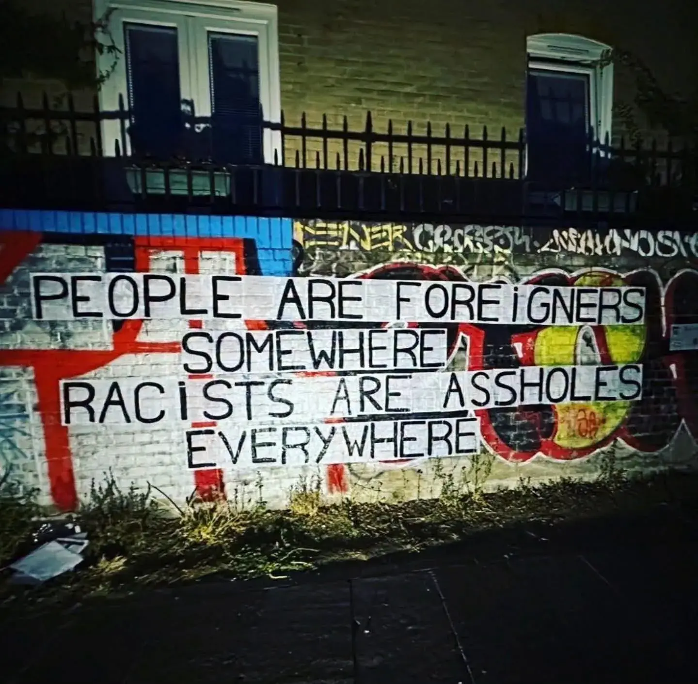 A photo of anti-racist street graffiti painted onto a brick wall beneath a residential railing. The text written in bold, uppercase lettering reads: "PEOPLE ARE FOREIGNERS SOMEWHERE. RACISTS ARE ASSHOLES EVERYWHERE