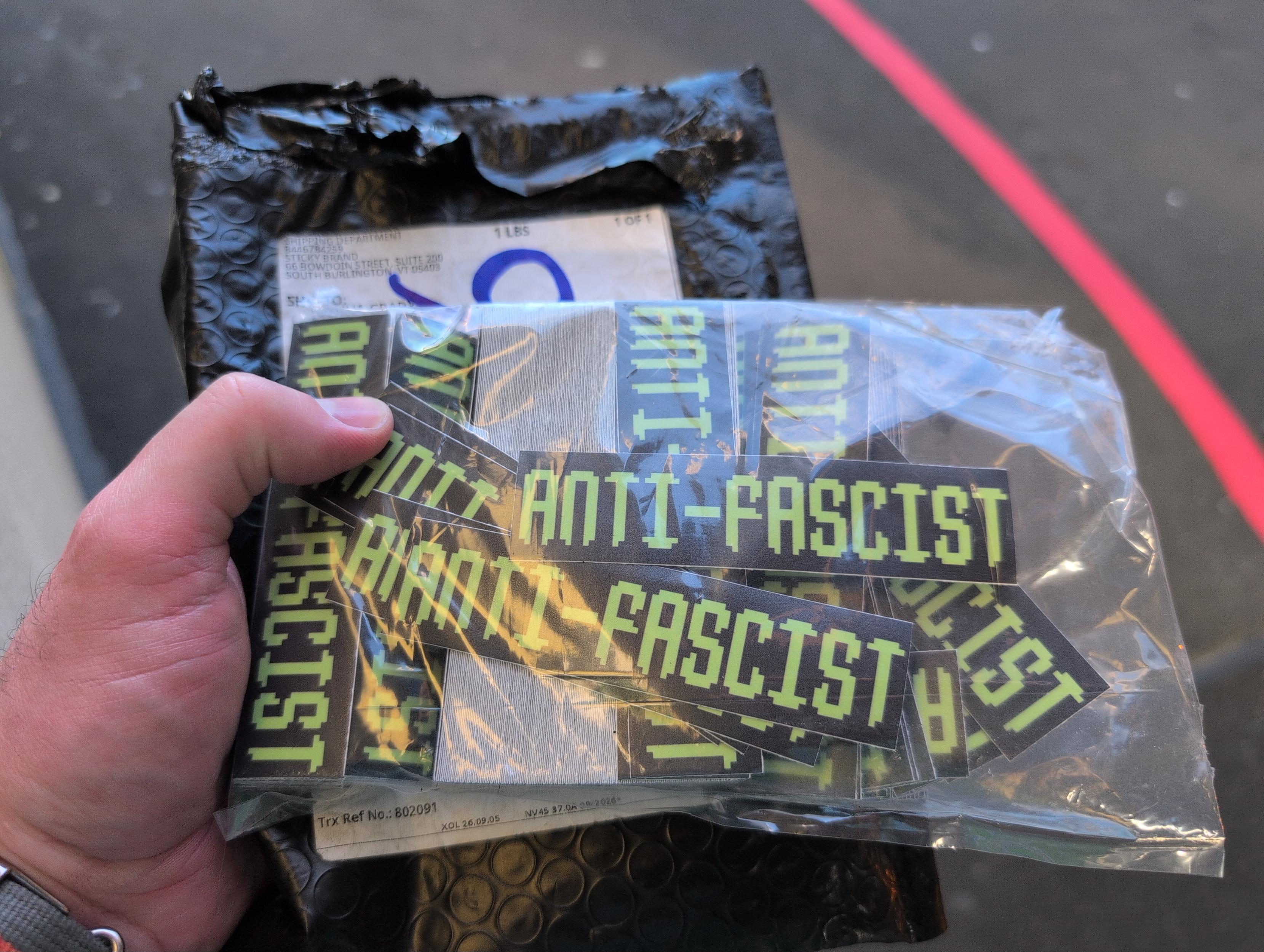 Me holding an open package of stickers that say "ANTI-FACIST" in an old-school video game-looking font.