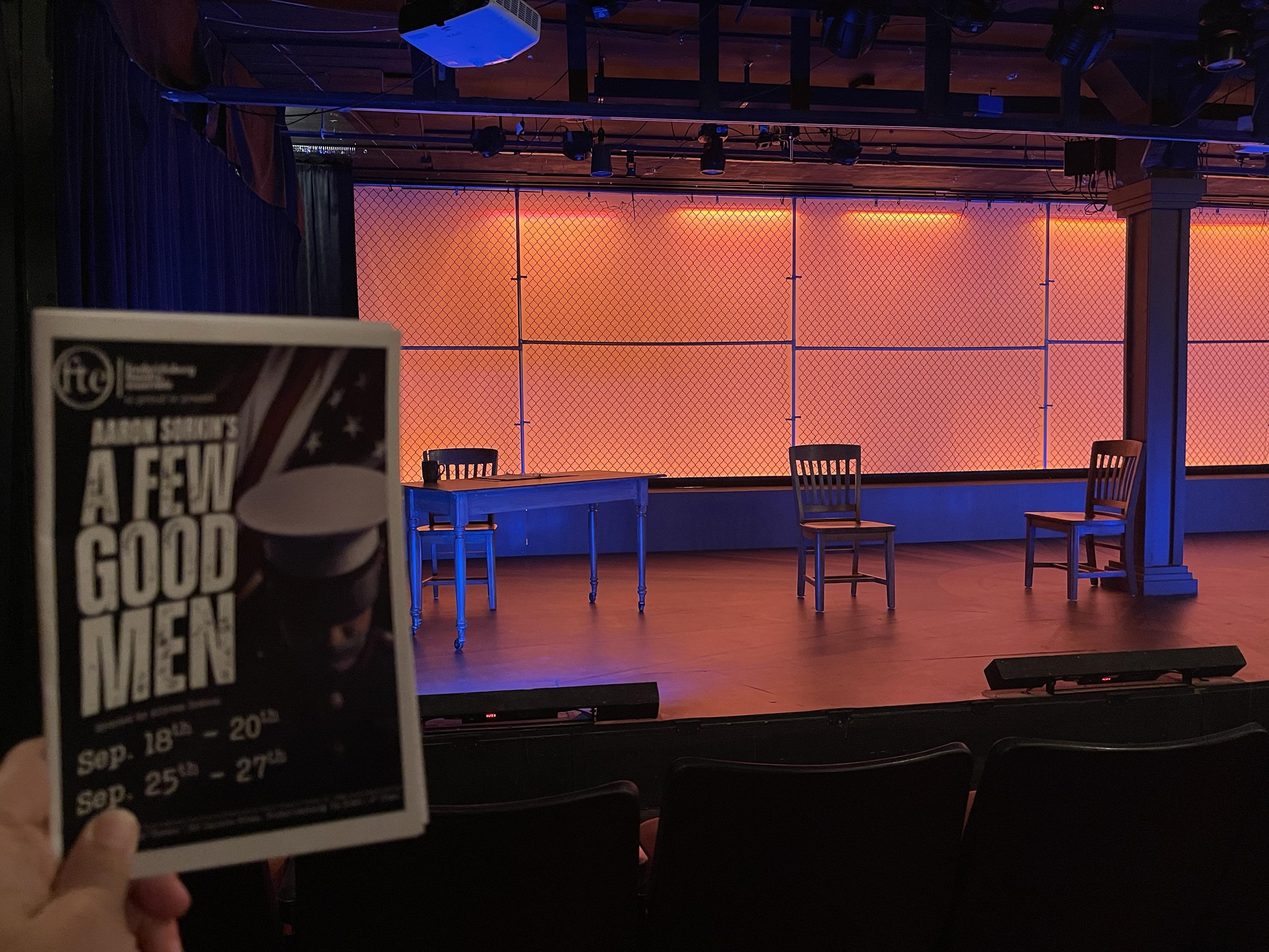 A playbill showing "Aaron Sorkin's A Few Good Men" held in front of a lit set prior to the play starting. It's a small, intimate theater where you're seated right up next to the stage. The stage is set simply and backlit ominously.