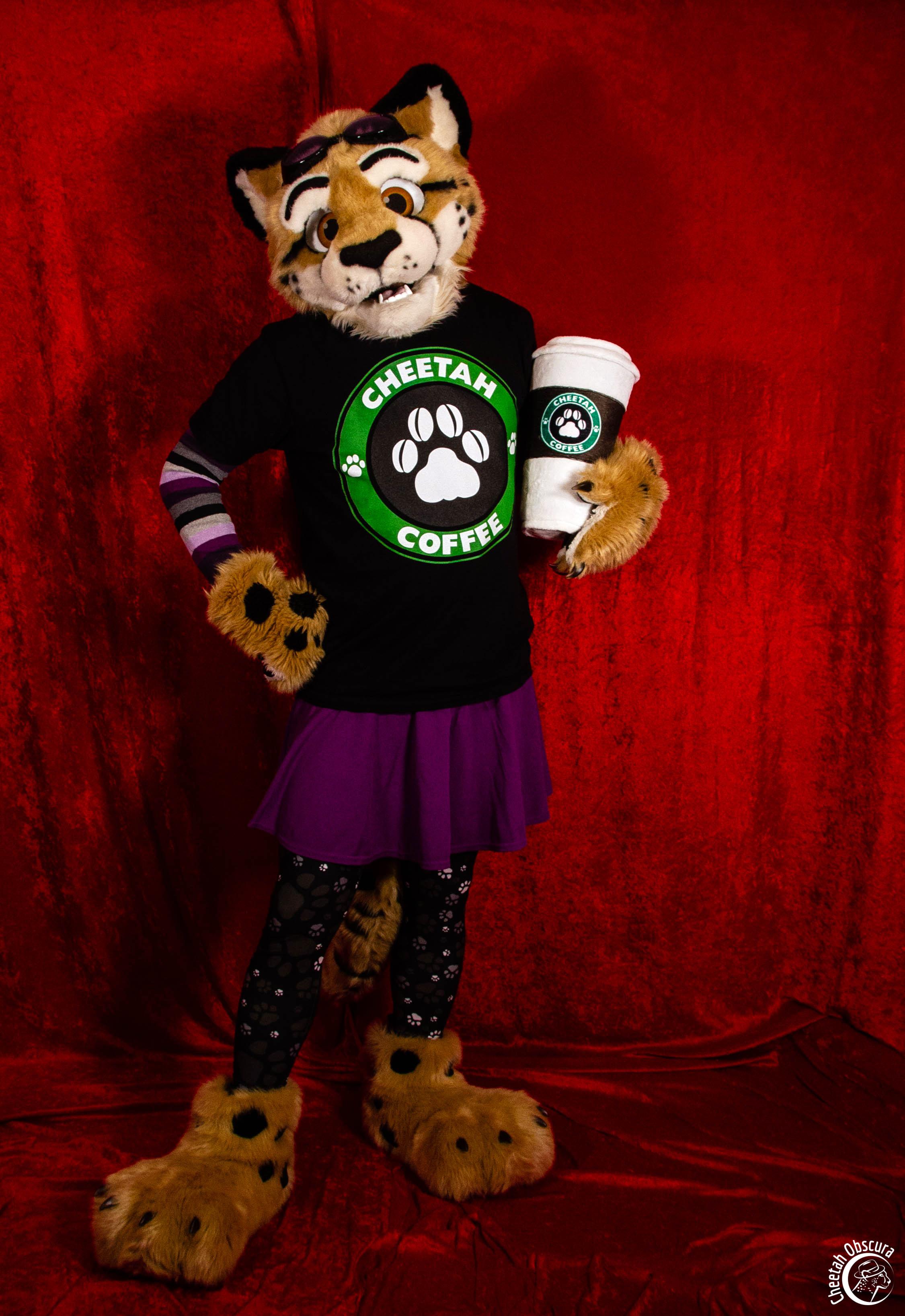 Fursuit photo of Obscura standing in front of a red background. They are wearing a black "Cheetah Coffee" shirt, purple skirt, paw print leggings, and asexual colors arm sleeves. They are holding a giant plush coffee cup with the "Cheetah Coffee" logo on it