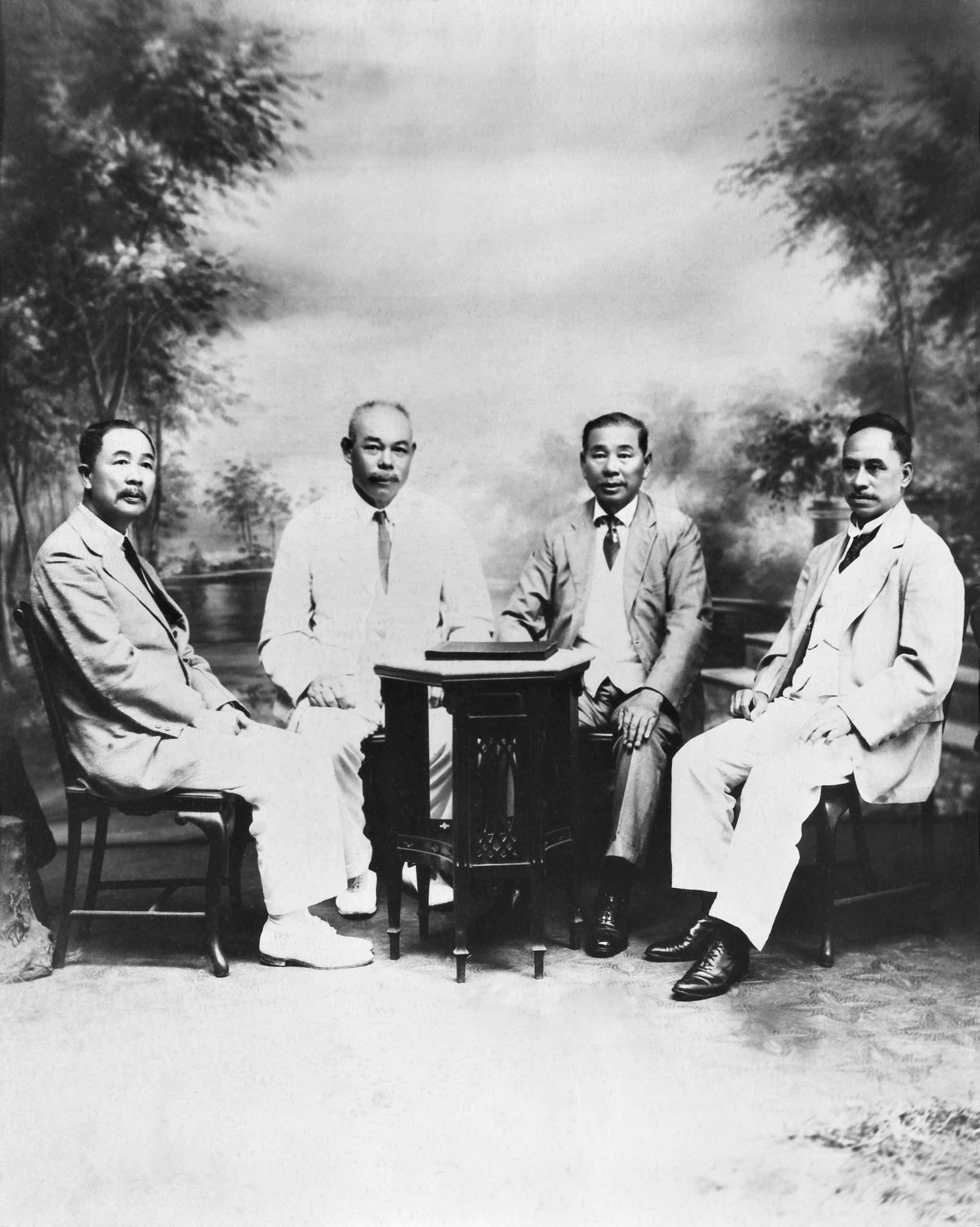 Very old black and white formal studio photo of four Chinese businessmen in light-coloured suits and dark ties sitting around a small table. A painted backdrop shows trees and a cloudy sky.