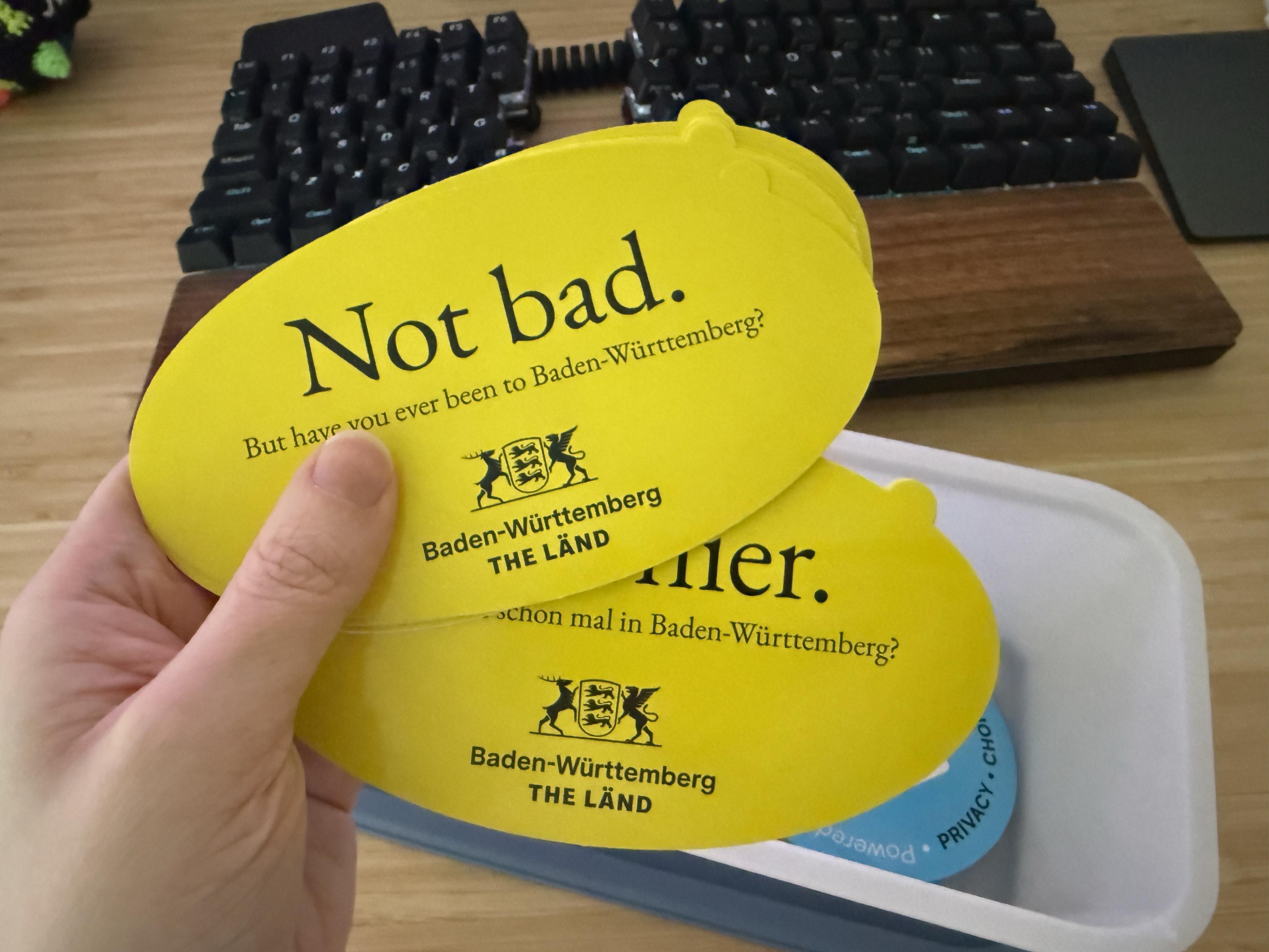 two giant elliptical stickers in my hand. they're yellow, and they say "Not bad. But have you ever been to Baden-Württemberg?"