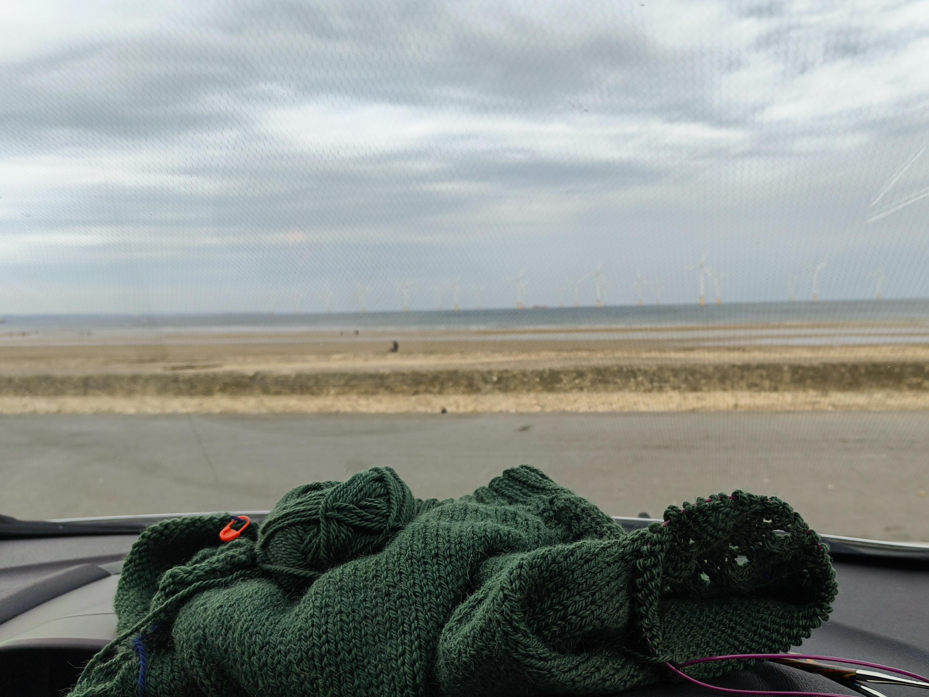 View through the windscreen of my parked car, with the beach and sea out of focus in the distance under a grey cloudy sky. You can also just about make out the windmills of the offshore wind farm. In the foreground, lying on top of the car dashboard, is a dark green knitted jumper with live sleeve stitches on a circular needle and the current ball of yarn nestled on top. From this angle the jumper looks like mostly stockinette although there's a peek of lace on the in-progress sleeve section.