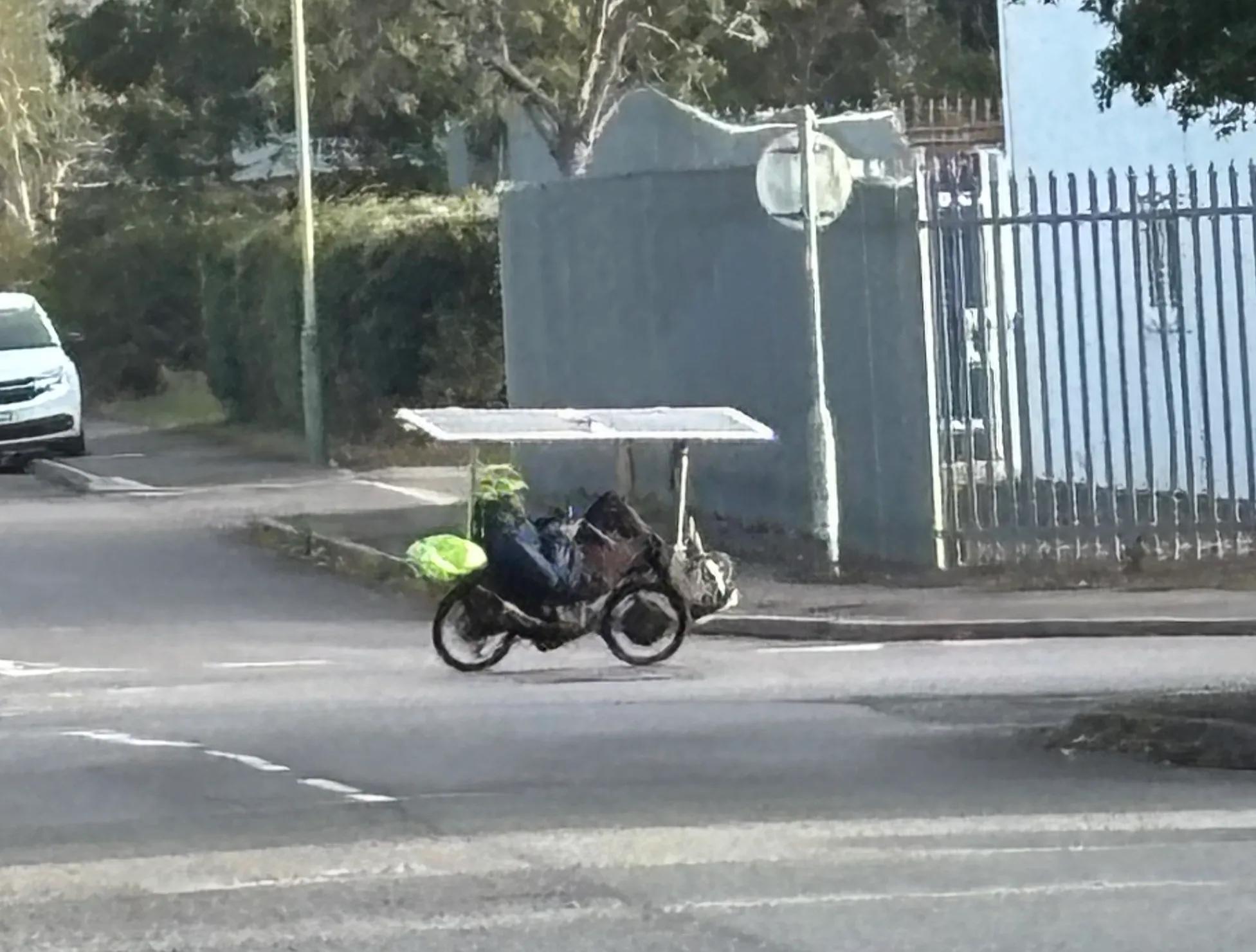 Industrial estate with a small grainy cyclist turning the corner, laying back on a recumbent bike, and two flexible solar panels making a roof over the top.