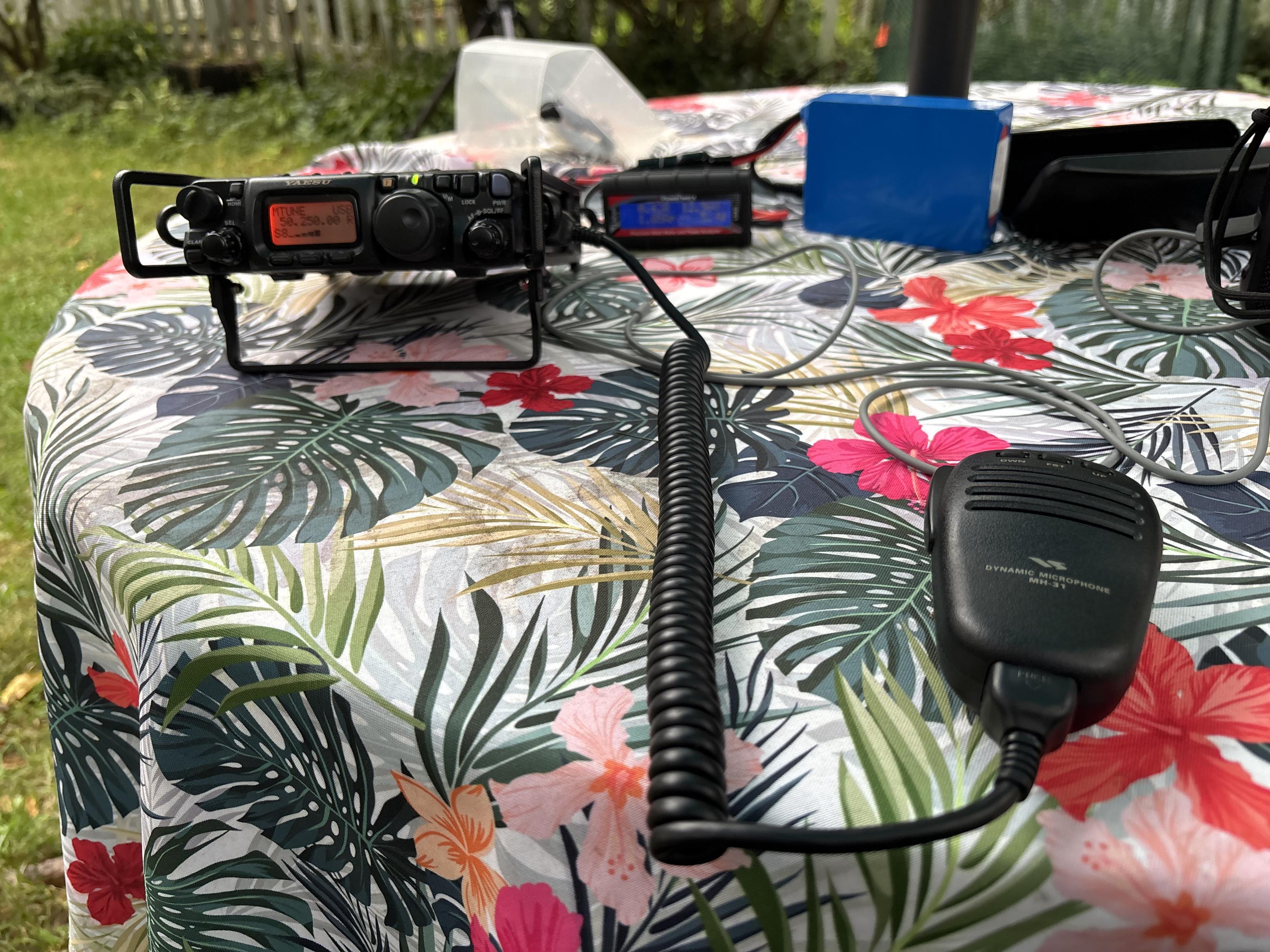Yaesu FT-818 all band/all mode QRP transceiver, microphone, and battery set up on table in back yard and connected to a homebrew square quad loop antenna