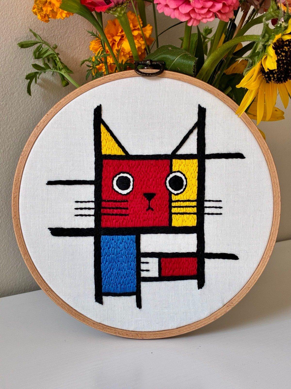 Handmade Meowdrian Embroidery. This embroidery is presented in a light wood display hoop, ready for hanging. It is inspired by the paintings of Dutch painter Piet Mondrian, adapting his geometric style to a portrait of a cat.