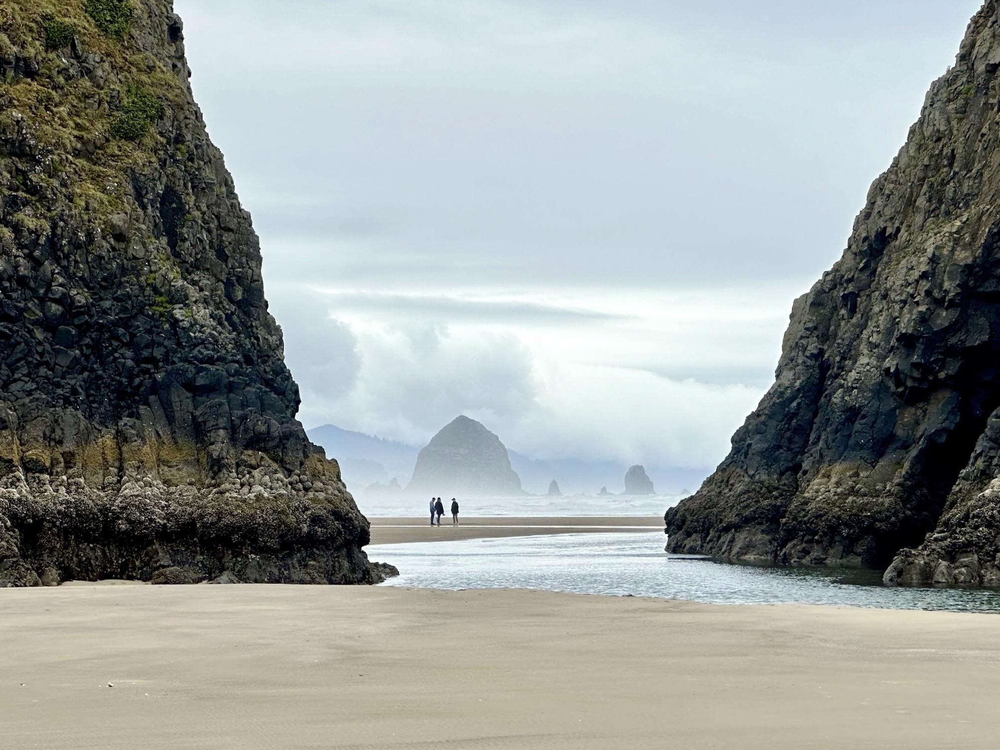 A sandy beach framed by towering rocky cliffs, with a few people walking in the distance. In the background, several sea stacks are visible under a cloudy sky. Taken at Chapman Point, Cannon Beach, OR, USA.