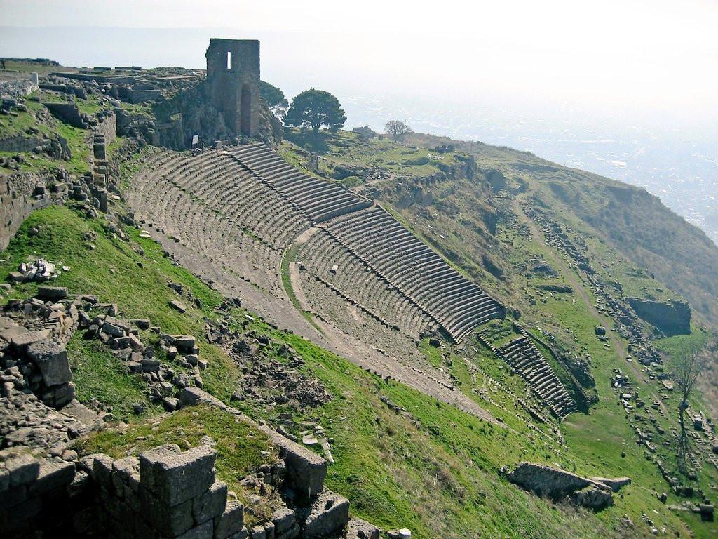 A full shot of the ruins of an ancient theatre, located on a hillside.  The theatre's steep tiered seating structure is clearly visible, constructed of light gray stone blocks, weathering evident in various tones. The hillside surrounding the theatre is covered with grass, creating a contrast between the man-made stone structures and the natural landscape. Sections of crumbling stone walls and foundations are scattered throughout the grassy slopes.
