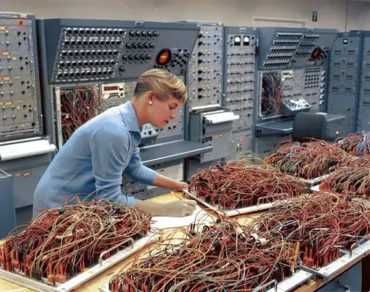 She is shown leaning over a desk, analyzing paperwork while surrounded by large analog computing consoles, which basically look like huge trays of spaghetti.