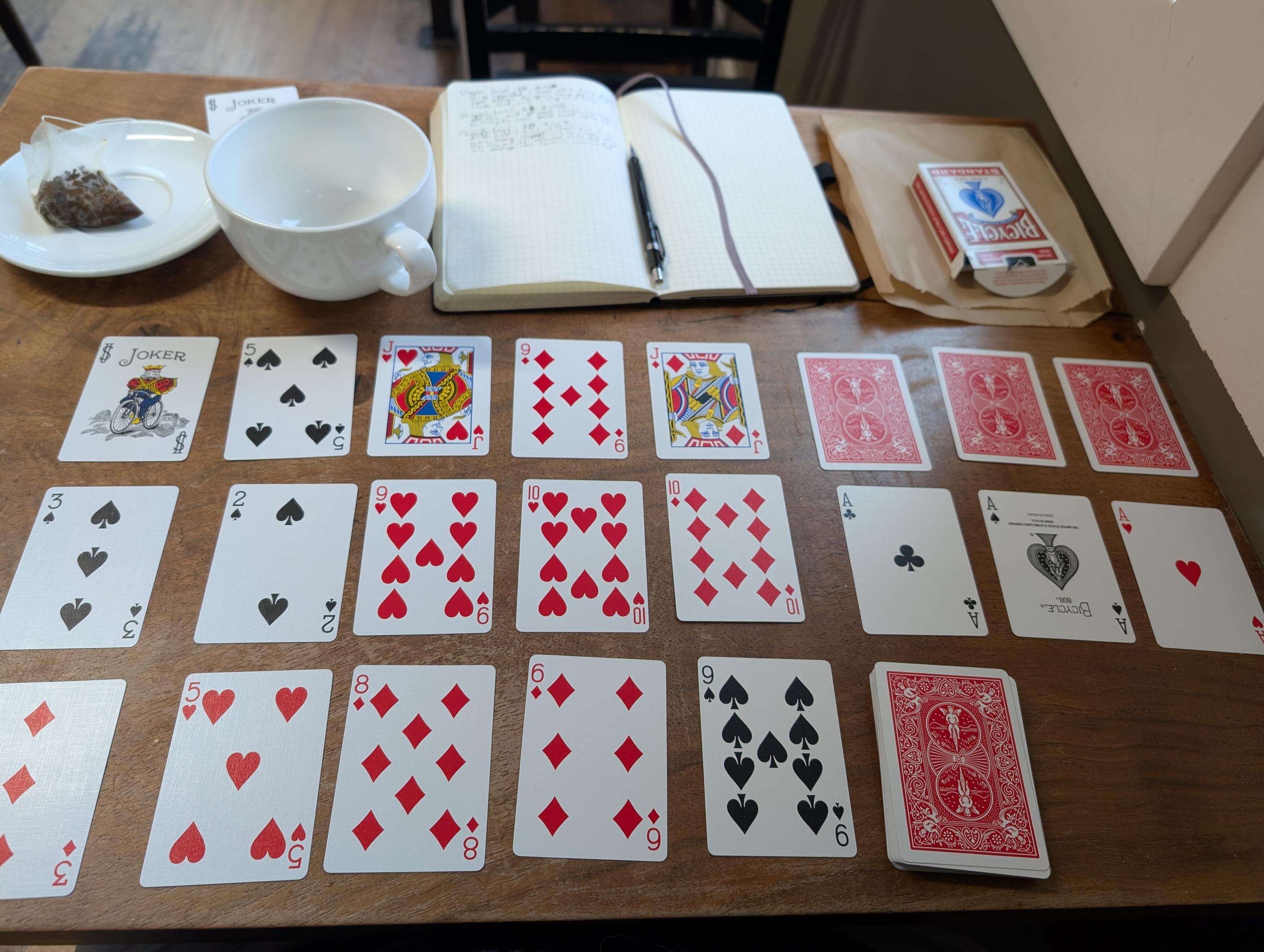 cards arranged on a coffee shop table in a large grid with some other stuff above it, like a cup, saucer, notebook, etc.