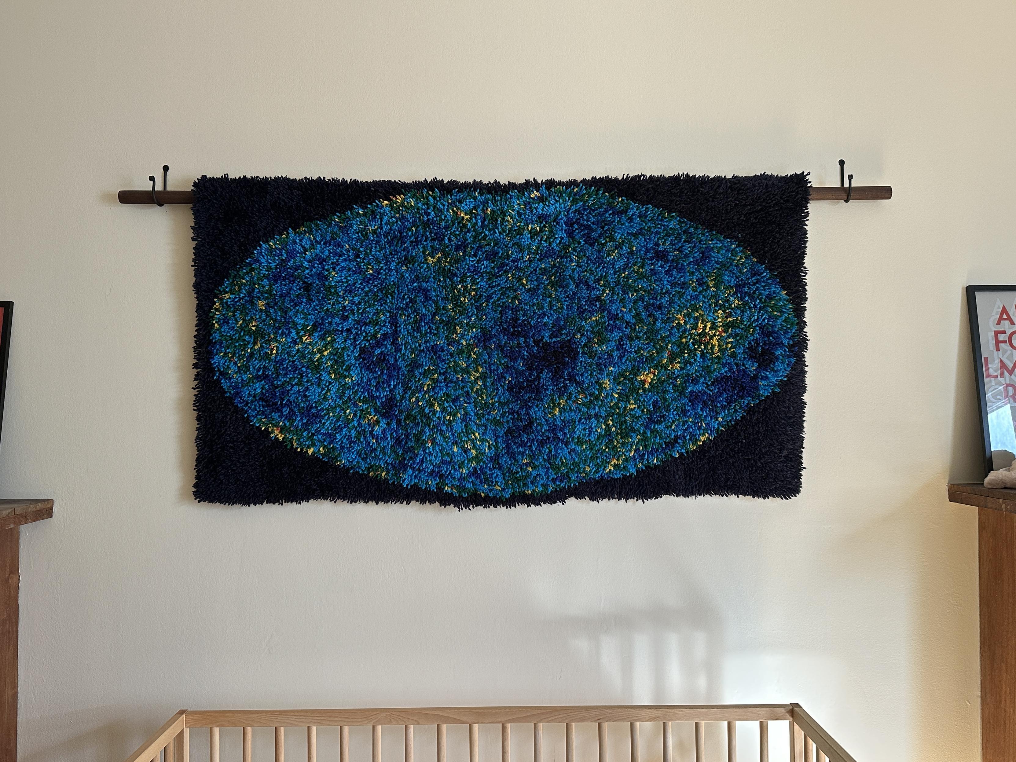 A homemade latch hook rug of the cosmic microwave background map, hung on a rod over a crib
