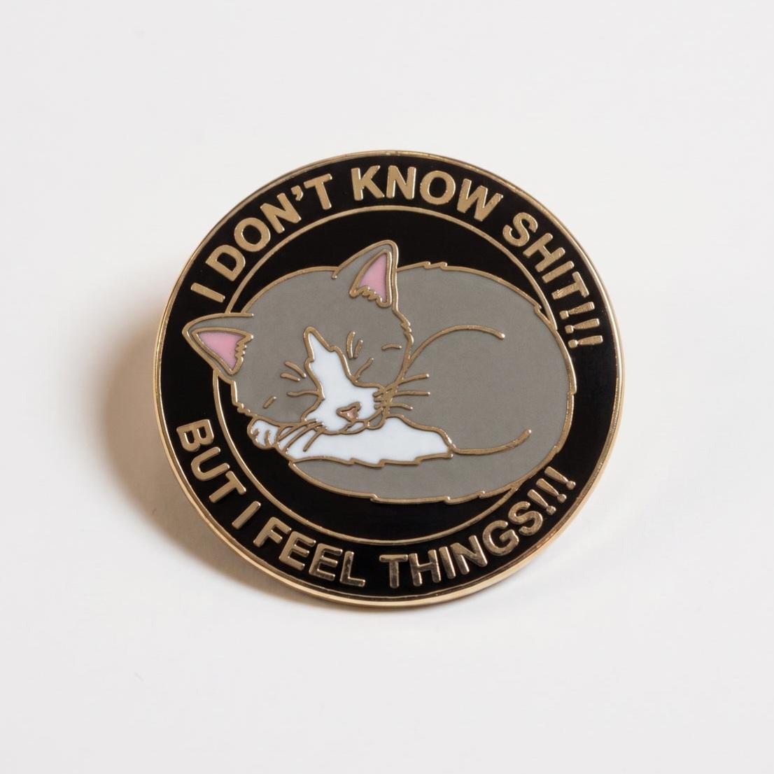 An enamel pin with a black circular border featuring a sleeping gray and white cat in the center. The text along the top curves to read "I DON'T KNOW SHIT!!!" and along the bottom reads "BUT I FEEL THINGS!!!".