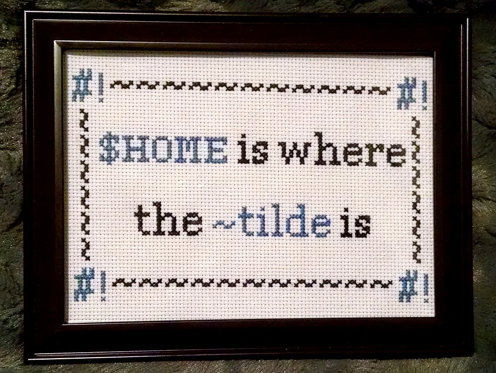 Framed cross-stitch that says "$HOME is where the ~tilde is." It has a border of tildes with #! shebangs in the corners.