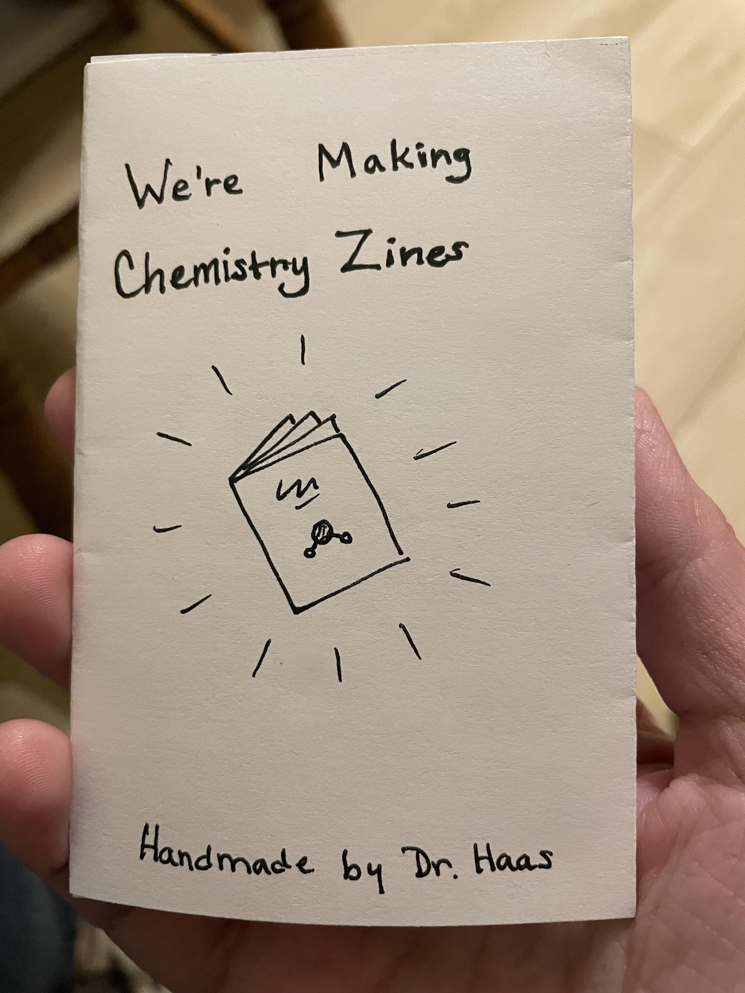 A little stapleless folded zine booklet titled "We're making chemistry zines" with a sketch of a zine with a molecule on it. The lettering is a little bit crooked and off-center. This zine proclaims itself handmade by Dr. Haas, and in the photo it's held in Dr. Haas's hand