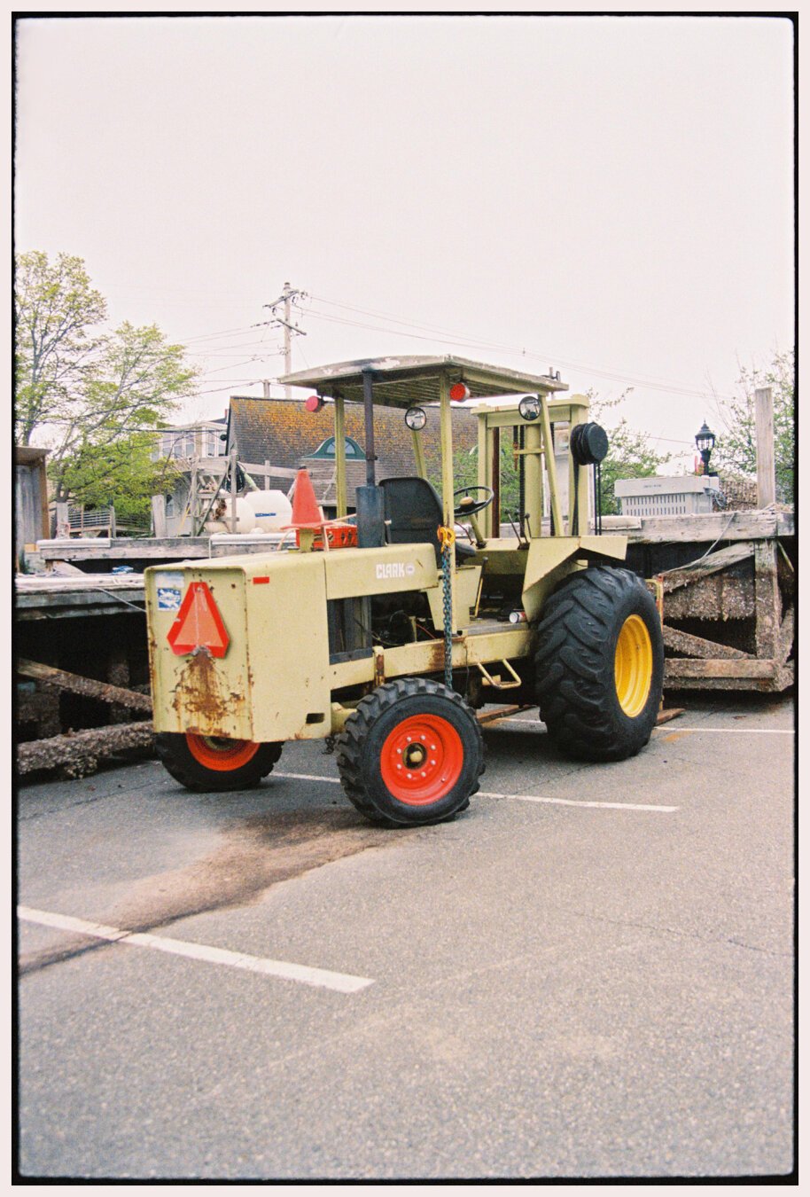 A photo taken with Kodak Portra 800 film in portrait orientation of an old forklift parked in front of the town’s dock floats which have been pulled out of the water for the winter and stored at the side of a parking lot. The forklift is an old-looking tractor-style model which is painted a pale avocado green. The rims of the large front wheels are bright yellow and those of the smaller rear wheels are bright orange. The sky in the background is hazy and overcast and appears a very pale gray.