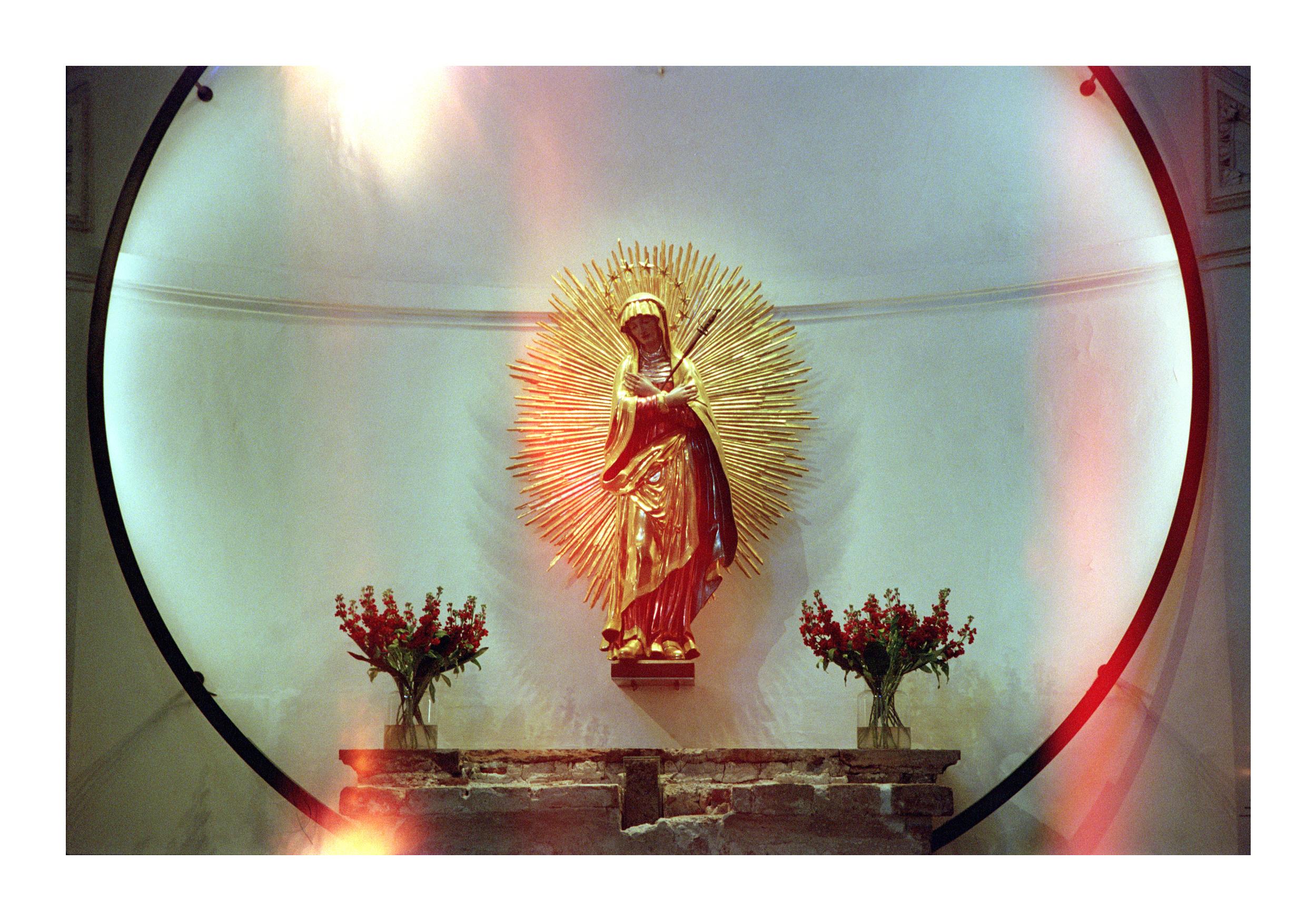 A film photograph capturing a golden statue of a robed figure with a sunburst halo, centrally placed on a weathered stone altar. The statue is flanked by two identical red flower arrangements in clear glass vases, symmetrically positioned on either side. Behind the altar, a large circular light flare in soft pink and red hues frames the scene, its edges slightly blurred. The film grain adds texture, and the warm, diffused light enhances the golden tones of the statue while casting subtle shadows. The composition balances the sacred subject with the accidental beauty of the light leak, creating a serene yet dynamic atmosphere.