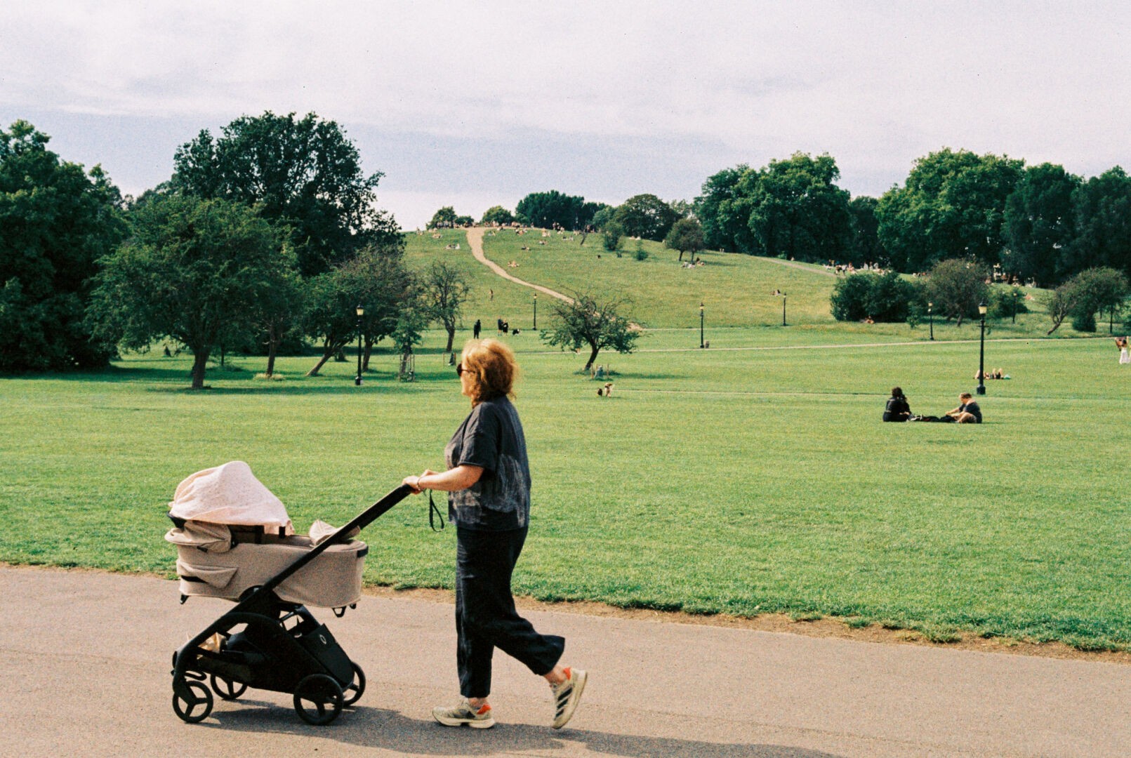 Woman pushing a stroller on a path in Primrose Hill, London, with a wide grassy hill and scattered trees in the background.