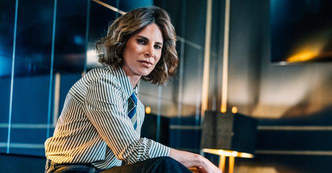 Jillian Michaels sits on the arm of a leather chair with one leg on the ground and the other resting on something off camera. She is wearing a tie, striped collared shirt and blue jeans.