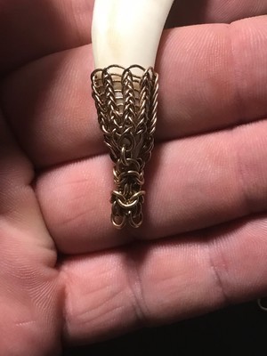 a hand holding a piece of boar's tusk partially wrapped in 20 gauge round bronze wire in a traditional ninth century wire weaving style transitioning smoothly into an intricate weave of wire rings in an equally interwoven three sided  chainmaille weave called Turkish Round