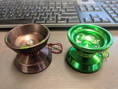 two yoyos on a brown desk. the left yoyo is aluminum, anodized in brown with green splashes. it has a polished aluminum spike on the axle hub, and a red/black string. the right yoyo is anodized green aluminum with a green/yellow string and a laser etched barracuda logo.