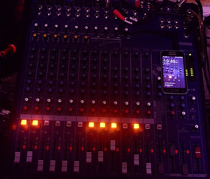 The superbly lighted photograph portrays a Yamaha audio mixing console with various potentiometers, faders, and switches. The console is predominantly dark blue with illuminated orange and red lights on the faders, indicating active channels. A smartphone is placed on the console, with a photograph of the same Yamaha mixing console! while displaying the time as 19:48. The console has multiple input and output jacks, with cables connected to it. The background is dimly lit, suggesting a low-light environment typical of a live performance or studio setting.

...the image is a rare glimpse into the alternate universe where Atmas rule the world.