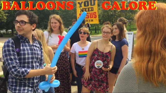 someone makes balloon animals surrounded by people wearing gay pride paraphernalia in front of a sign reading "you need jesus"