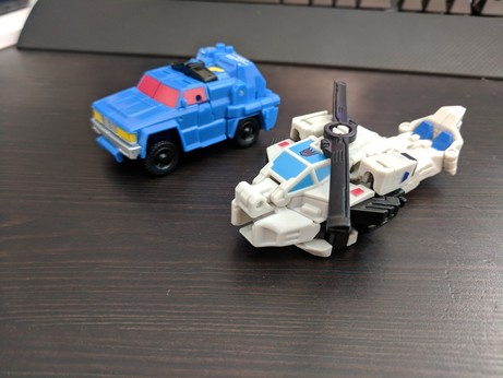 Transformers Generations Power of the Primes Legends figures Roadtrap and Battleslash. Roadtrap is a blue SUV with pink windows. Battleslash is a white helicoptor with light blue windows.