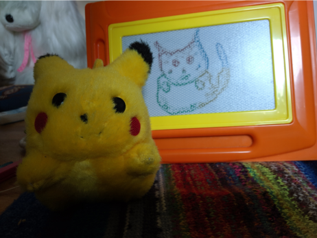 a pluche Pikachu sitting in front of an etch-a-sketch drawing of him