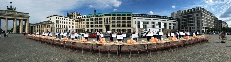 Eine lange Tafel, festlich gedeckt, steht auf dem Pariser Platz, dahinter eine Reihe von Menschen mit Schildern, auf denen ihr Beruf steht (z.B. "Hochzeits-Photograph") oder ähnliches. Links im Bild das Brandenburger Tor, rechts gibt jemand ein Fernseh-Interview.
