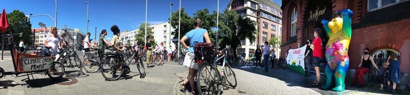 Menschen vor dem Rathaus Lichtenberg, viele mit Fahrrädern. Rechts ein "Buddy Bear", daneben Menschen mit einem Banner "Klimaschutz". An einem Lastenrad ganz links ein Schild "Climate over profit".