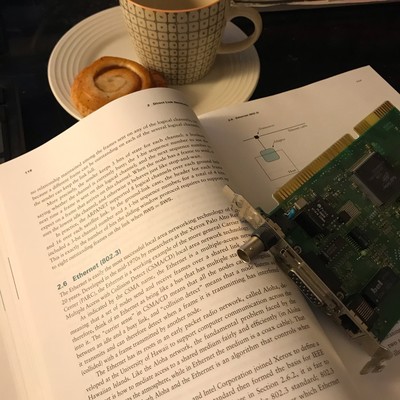Open book with text about ethernet and an old network card on top (3com EtherLink III). Tea and cinnamon bun in the background. 