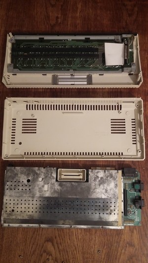 An opened Atari 600XL, 3 parts on a table: the keyboard, the ground plastics, and the actual board which is still closed with a somewhat dirty metal case.