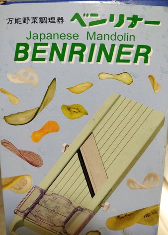 the box for a mandolin, BENRINER brand, featuring paper thin slices of vegetables and fruits raining down against a light blue background. 