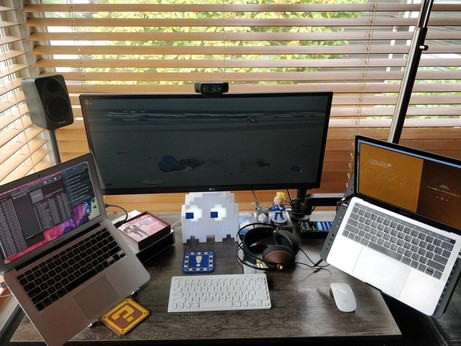 a small work desk in a corner with two laptops and a widescreen monitor. fancy headphones and a flashing plastic pacman ghost adorn the desktop. 